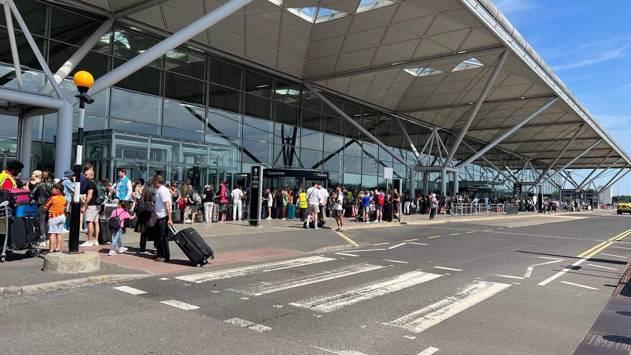 The outside of the departures area at Stansted Airport, with groups and queues of people outside the terminal building beside the dropping off bays on the road