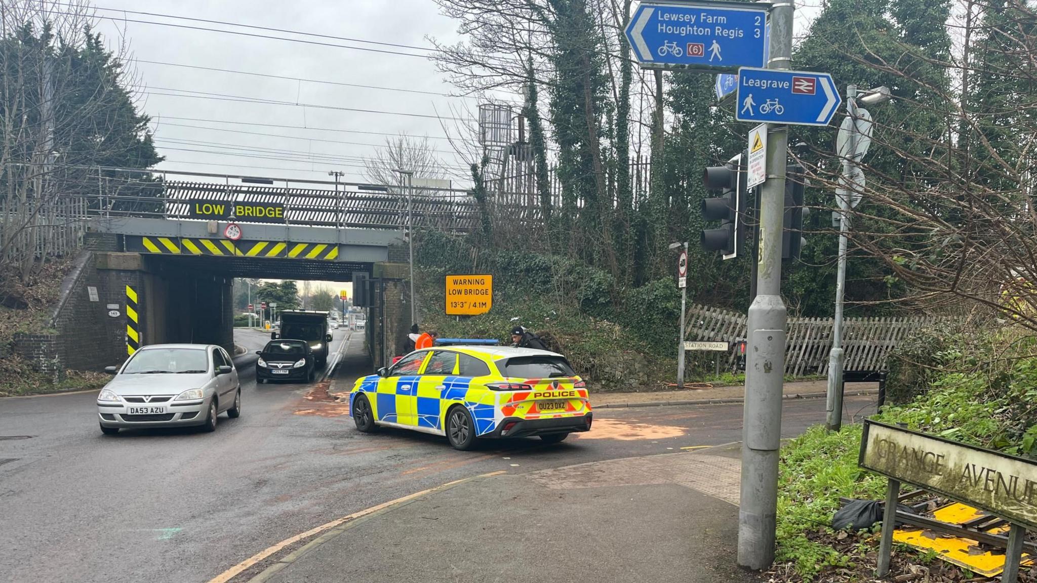 A police car blocks the entrance to Station Road. Cars pass under a railway bridge which has a "LOW BRIDGE" sign and yellow and black warning chevrons on it. A roadsign for Grange Avenue is in the foreground