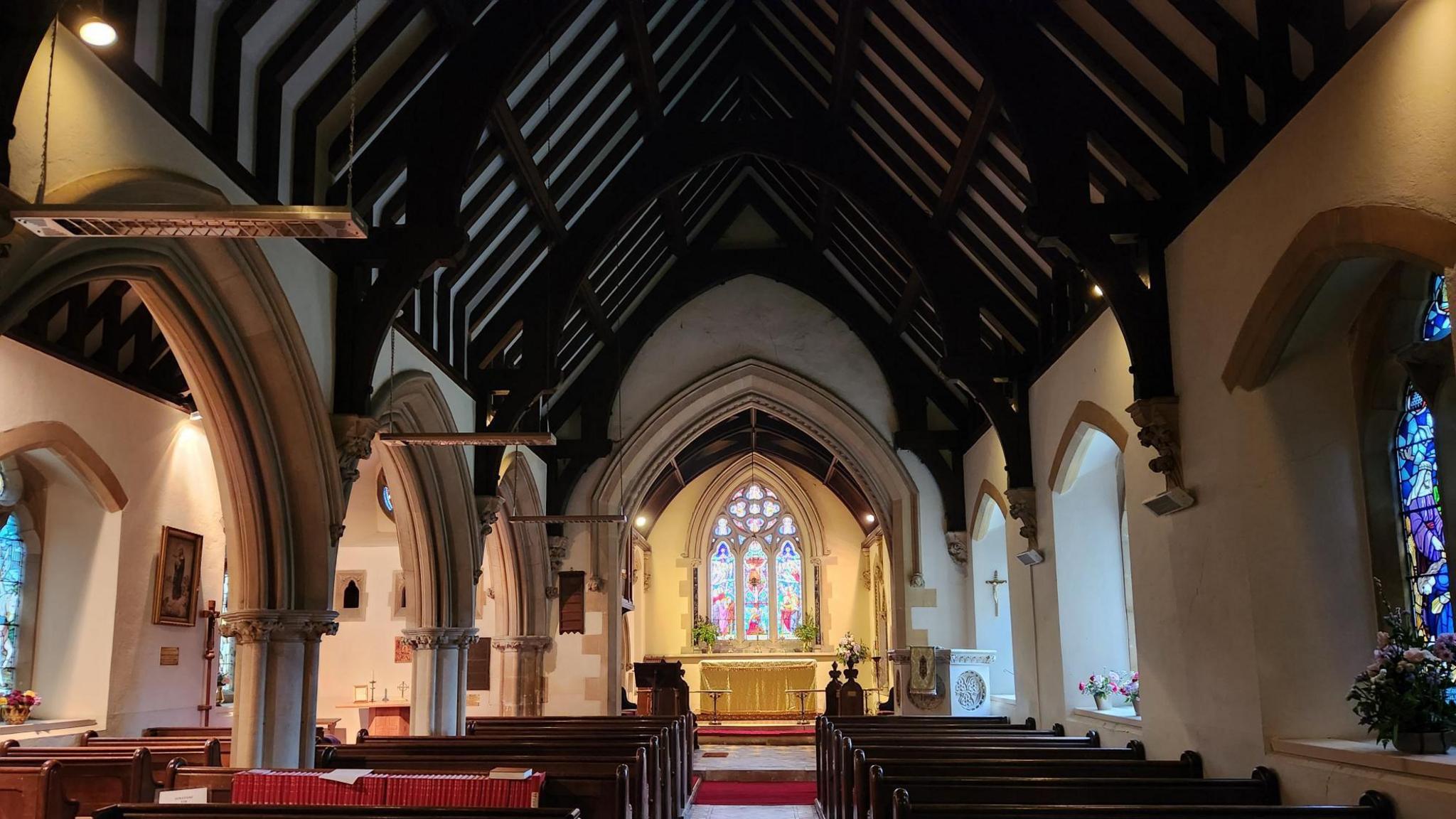 Inside of a church, looking down towards the altar which has a stained window behind it. The pitched roof has large wooden beams and there are large arches to the left next to pews