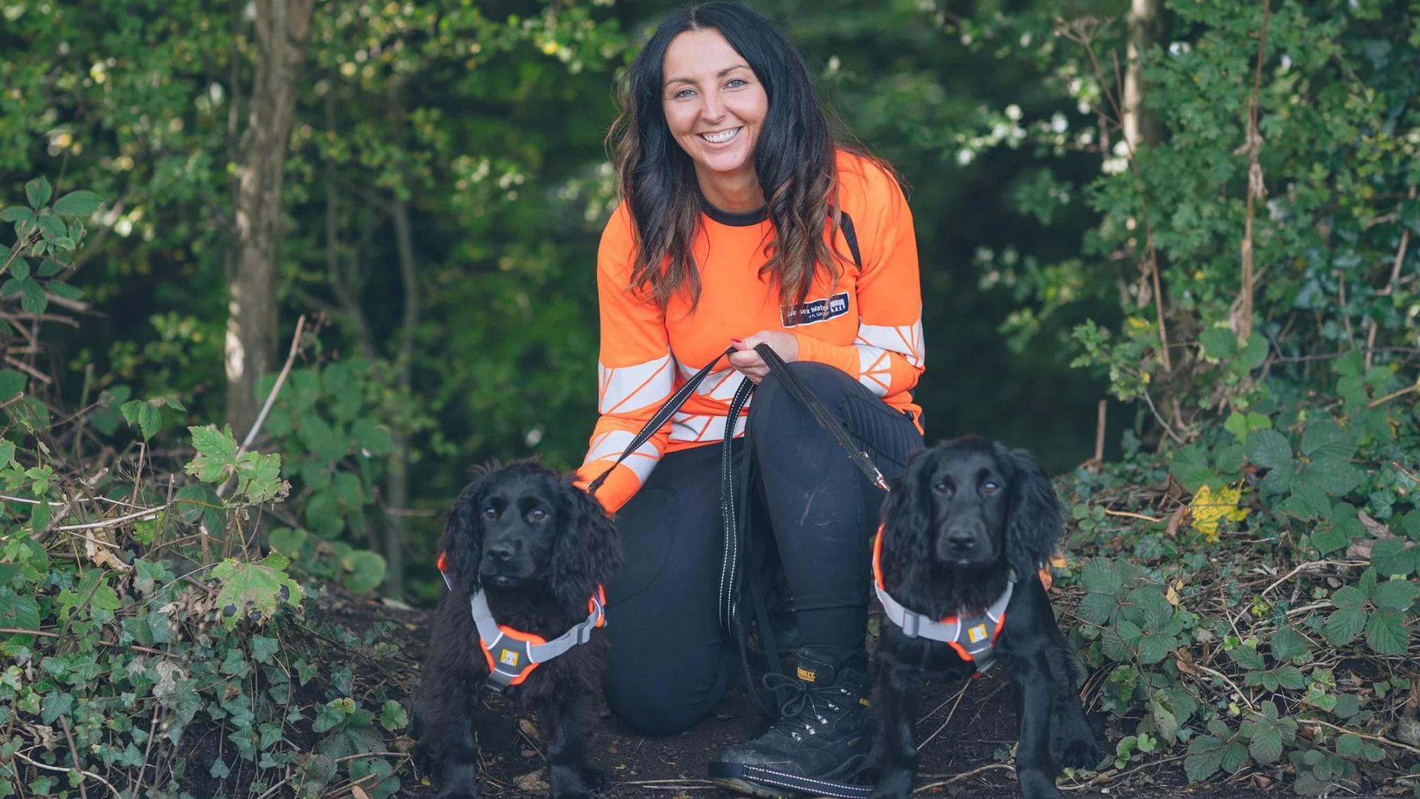 A woman wearing a high-vis orange shirt is sitting on the ground in the woods with two black dogs on a lead.