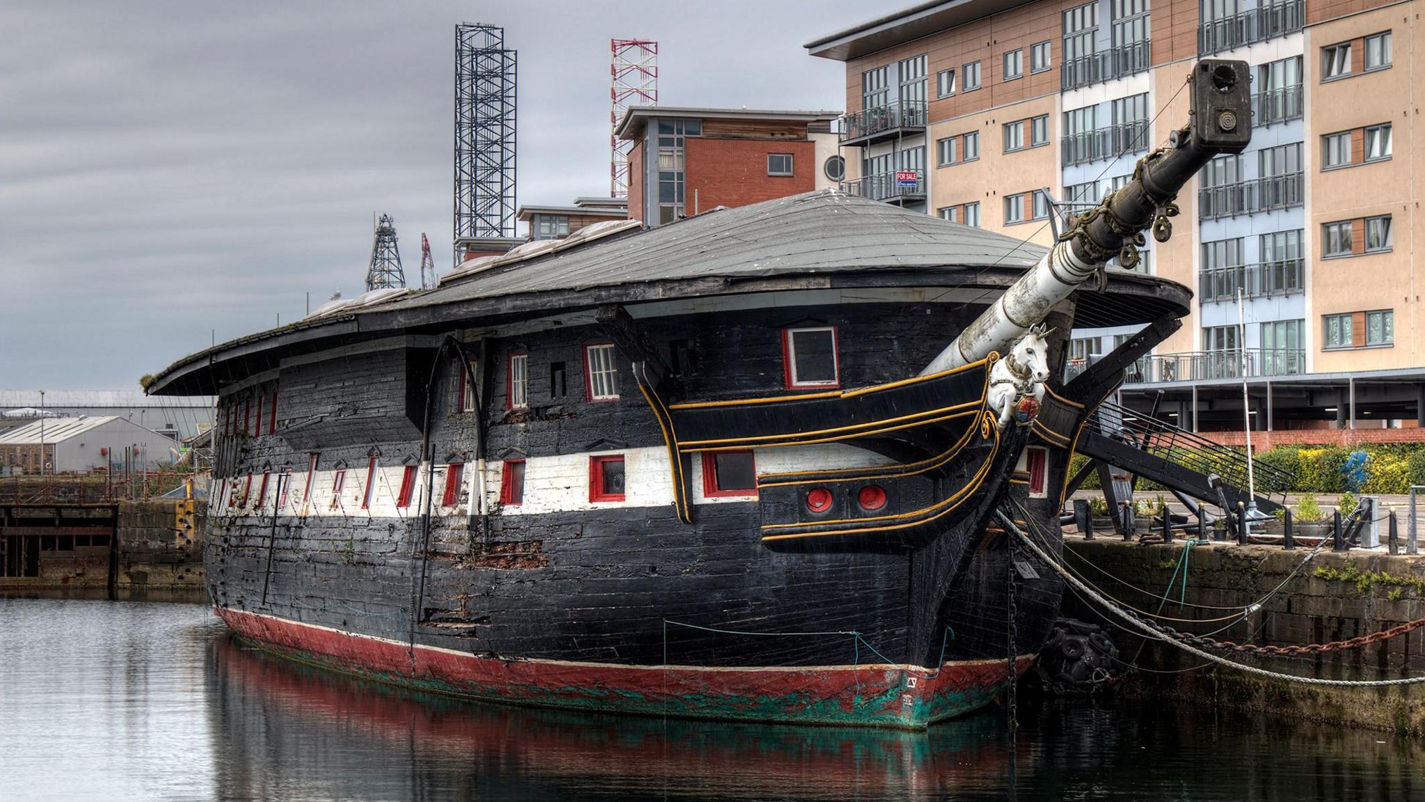 HMS Unicorn is the hulk of a 19th century timber sailing ship but has no rigging or sails. The hull is black and white striped with red window frames. It has an odd felt roof like a house. It is rather worn looking and stands in the water in front of some modern flats and a crane. The sky is grey.