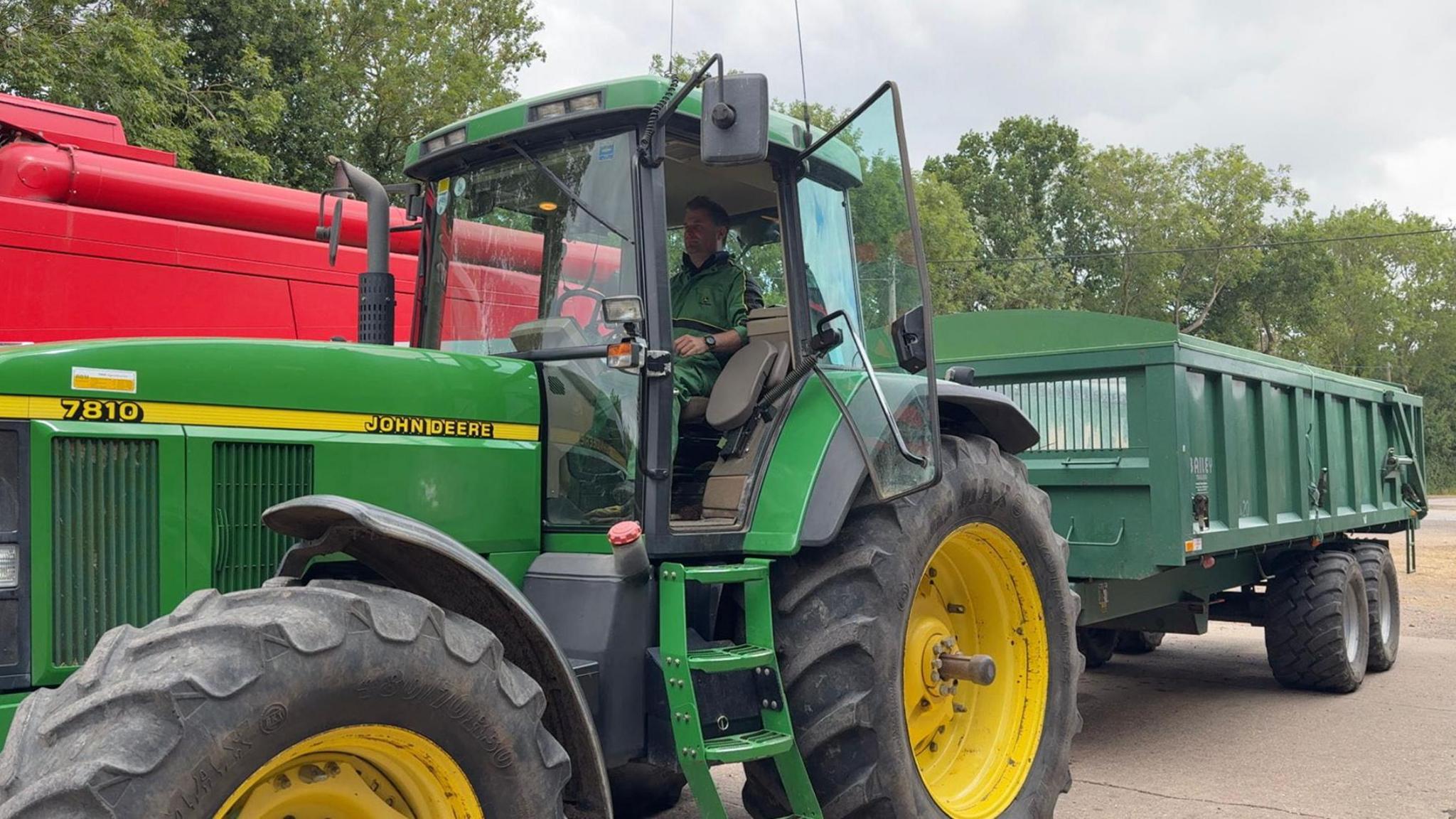 The picture shows a man sitting in a stationary, green tractor. It has huge wheels that are yellow in the middle. The tractor is pulling a green trailer.
