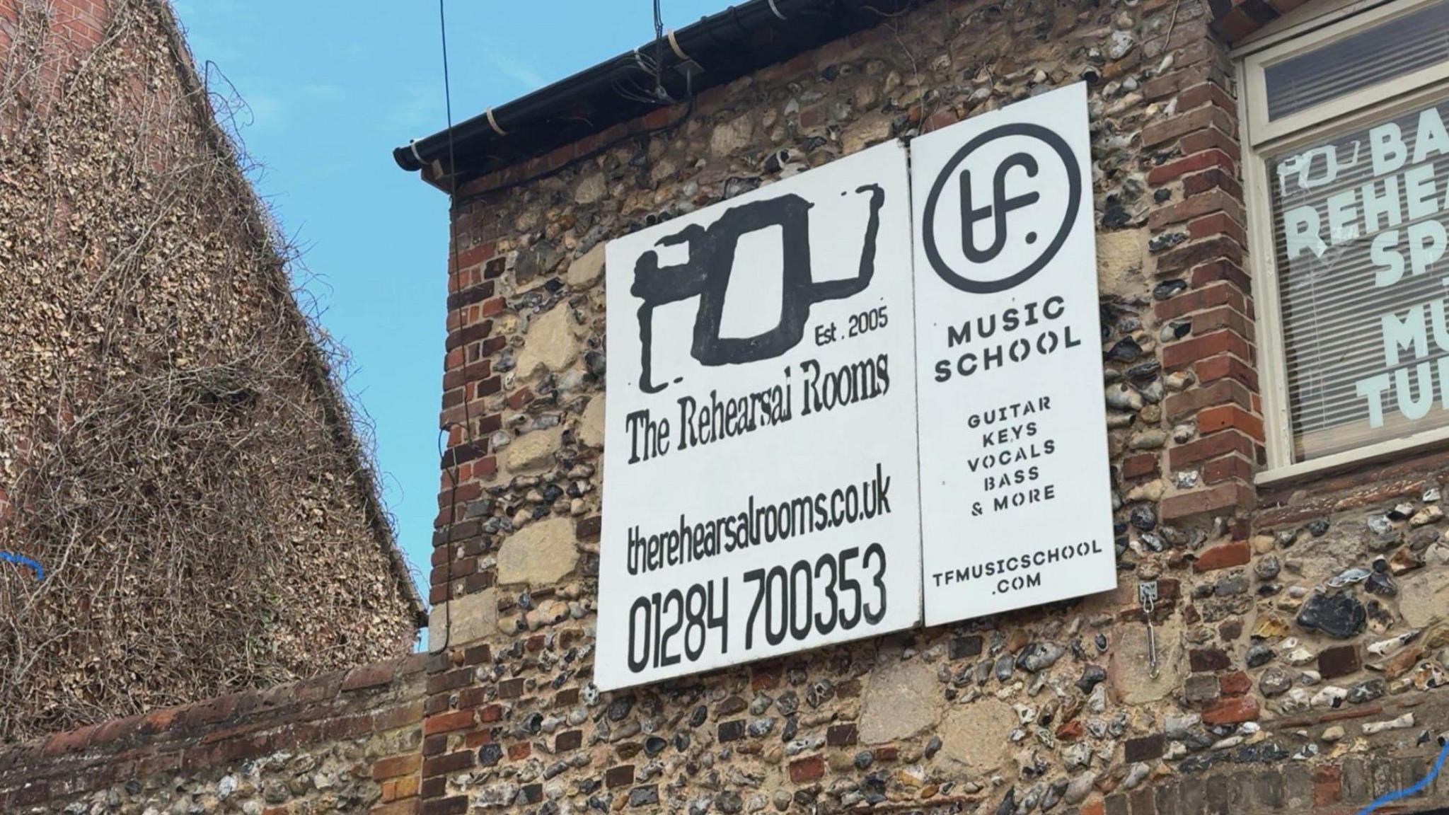 The top of a brick and stone building on which there is a large white and black sign for The Rehearsal Rooms music studio.