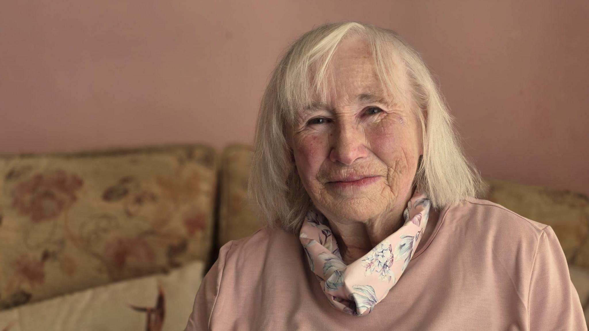A woman sat in a pink room on a beige floral sofa smiling.