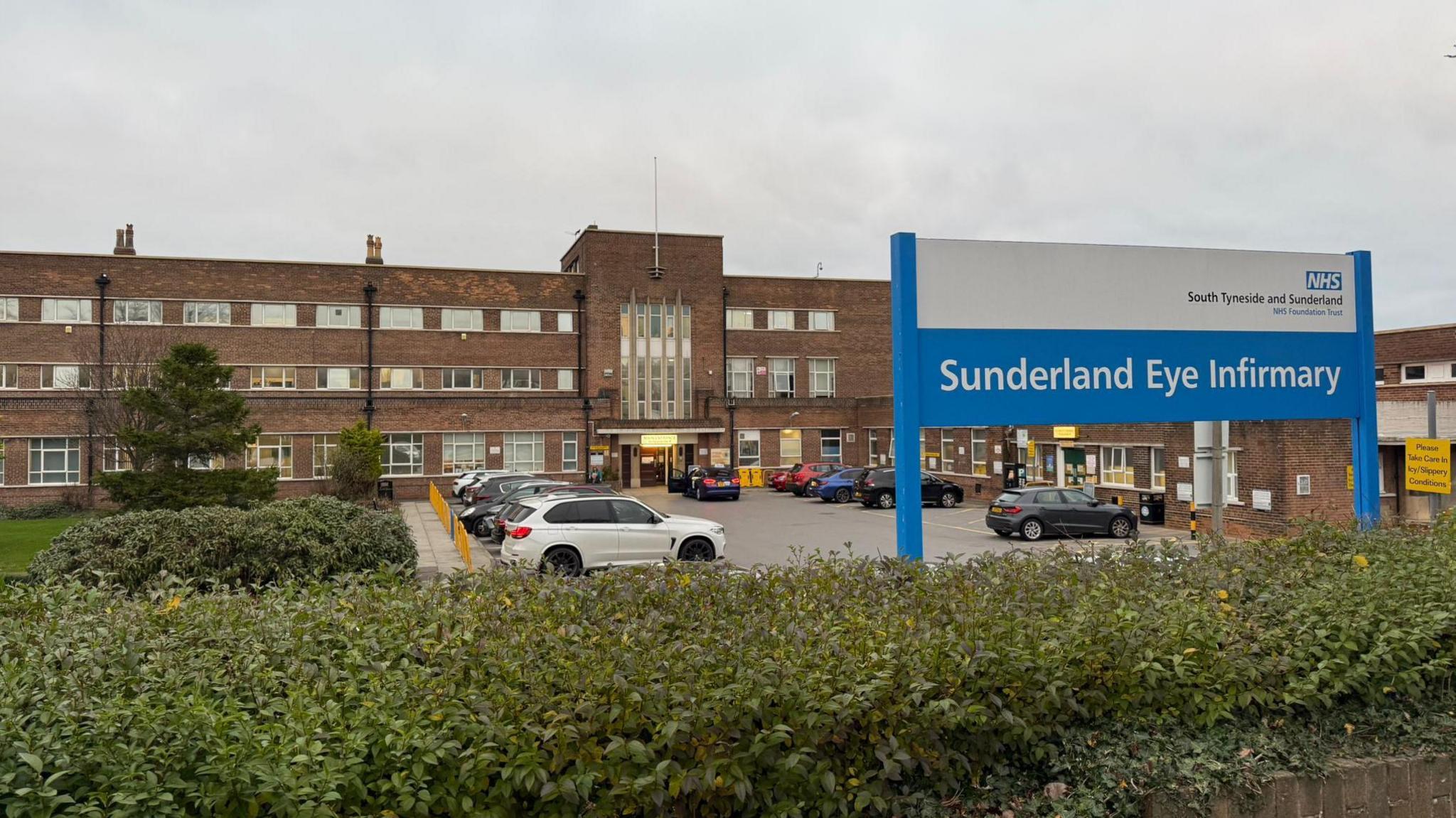 A white and blue NHS hospital sign saying Sunderland Eye Infirmary in front of the dated hospital building, which contains a brick façade.