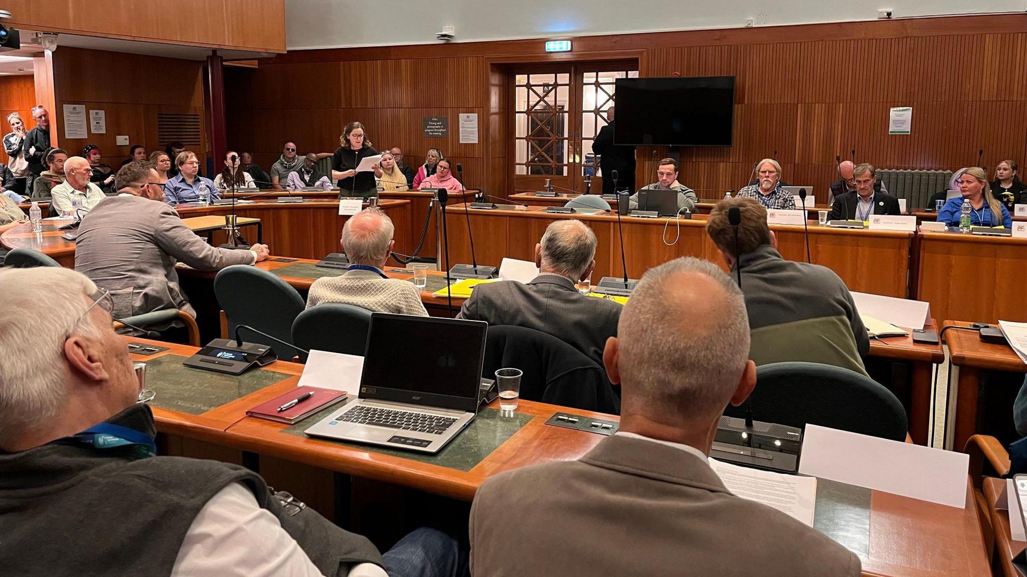 A group of councillors sit in the dark wood-panelled council chamber, watching on as a member of the public gives a speech read from a piece of paper. Members of the public can also be seen watching proceedings. 