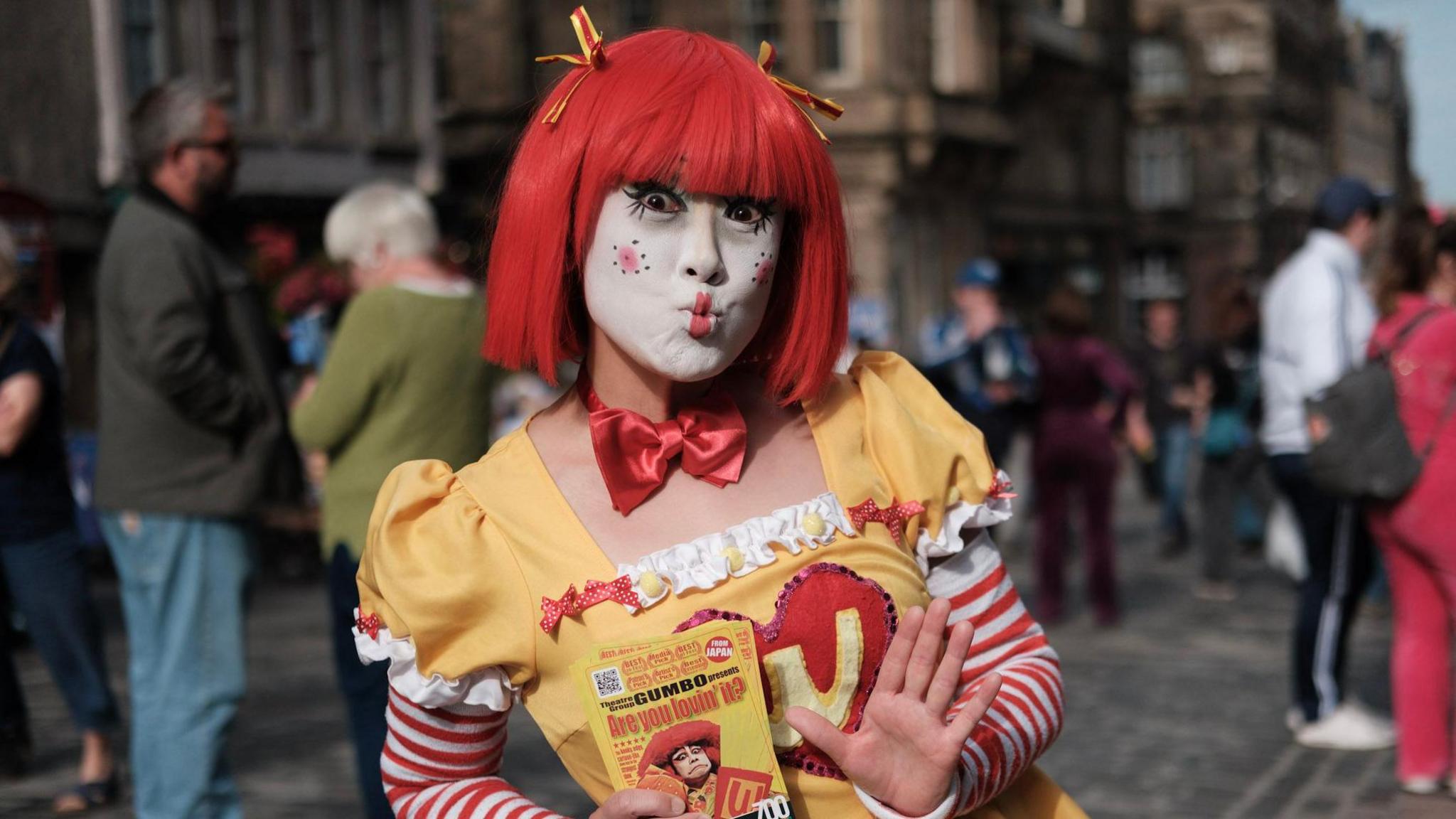 A doll-like clown in a yellow and red dress, bright red wig and doll clown make-up poses with a kiss while holding flyers advertising her show