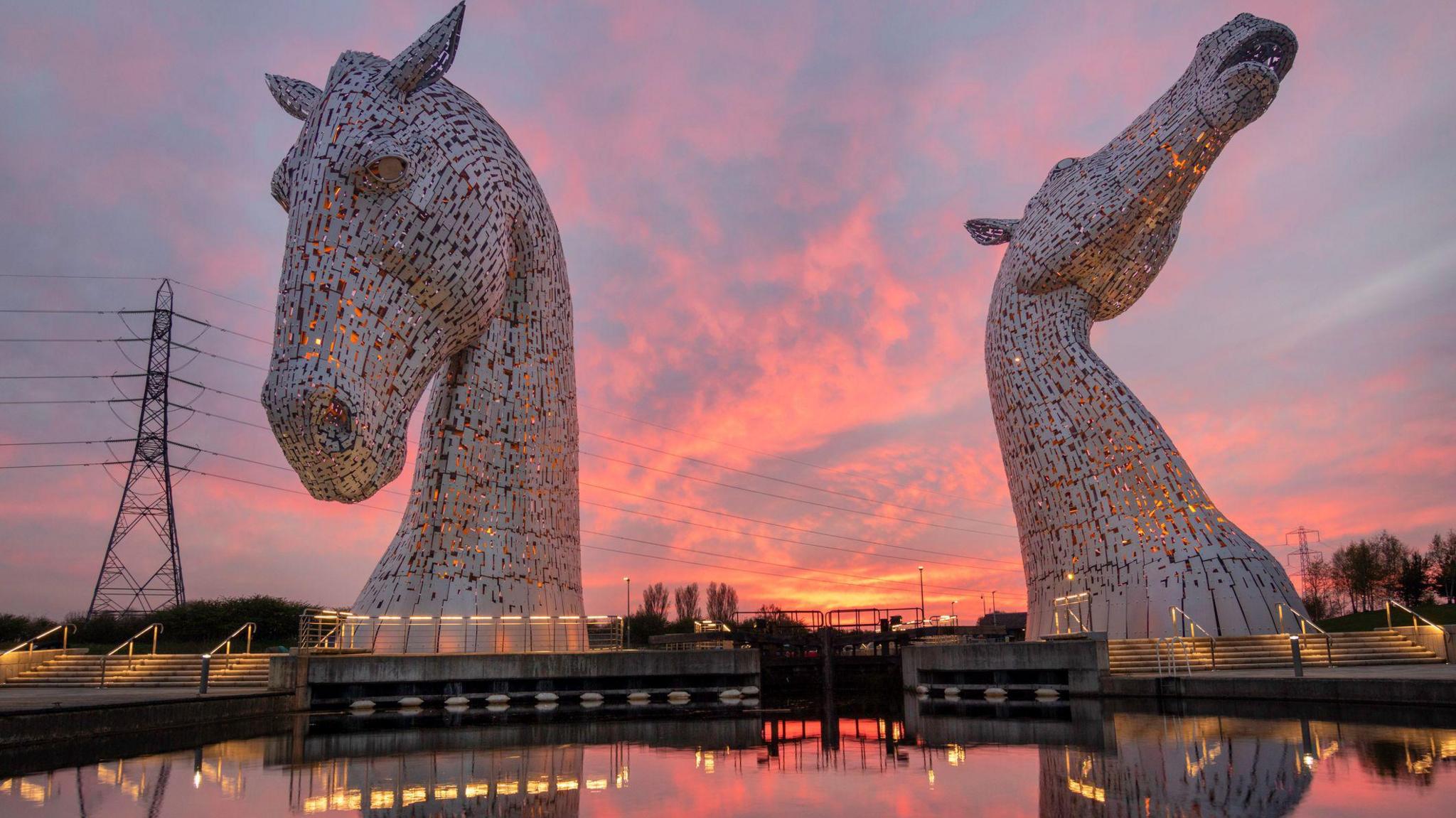 Two horses heads made from metal sit on the water with a beautiful pink sunset in the background.