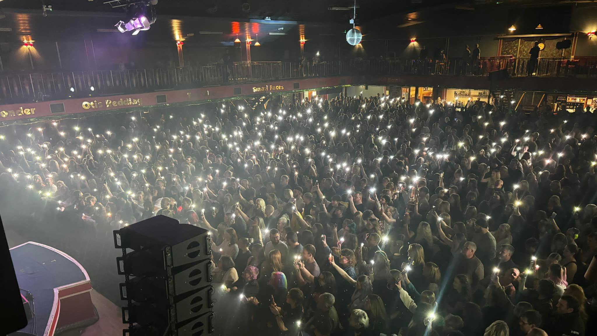 A crowd watching the stage (out of view to the left) are holding up lights on their mobile phones in front of Scouting for Girls