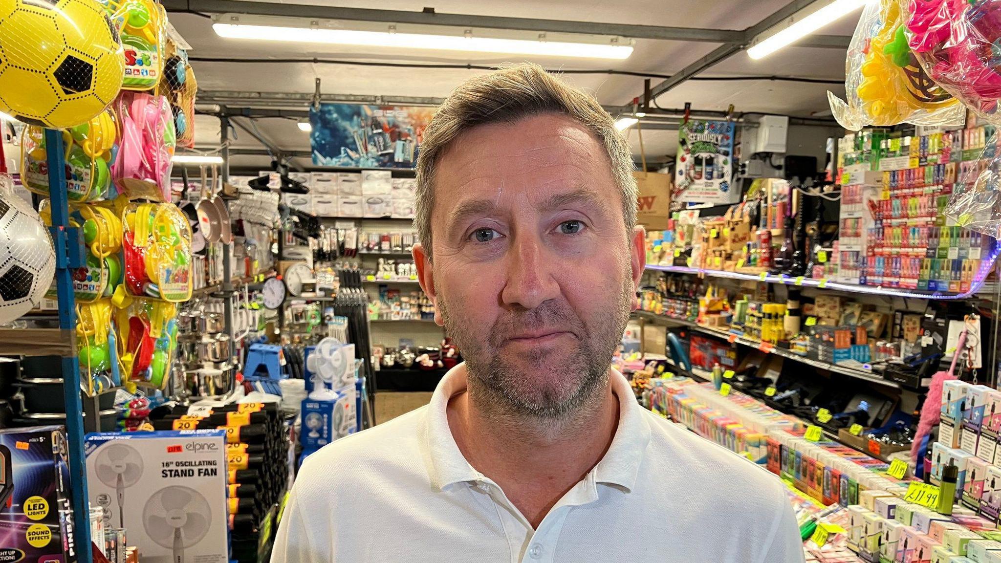 A picture of a man in front of the market stall he owns. It sells a wide variety of goods, and he is wearing a white polo shirt. He has short hair and a goattee beard.