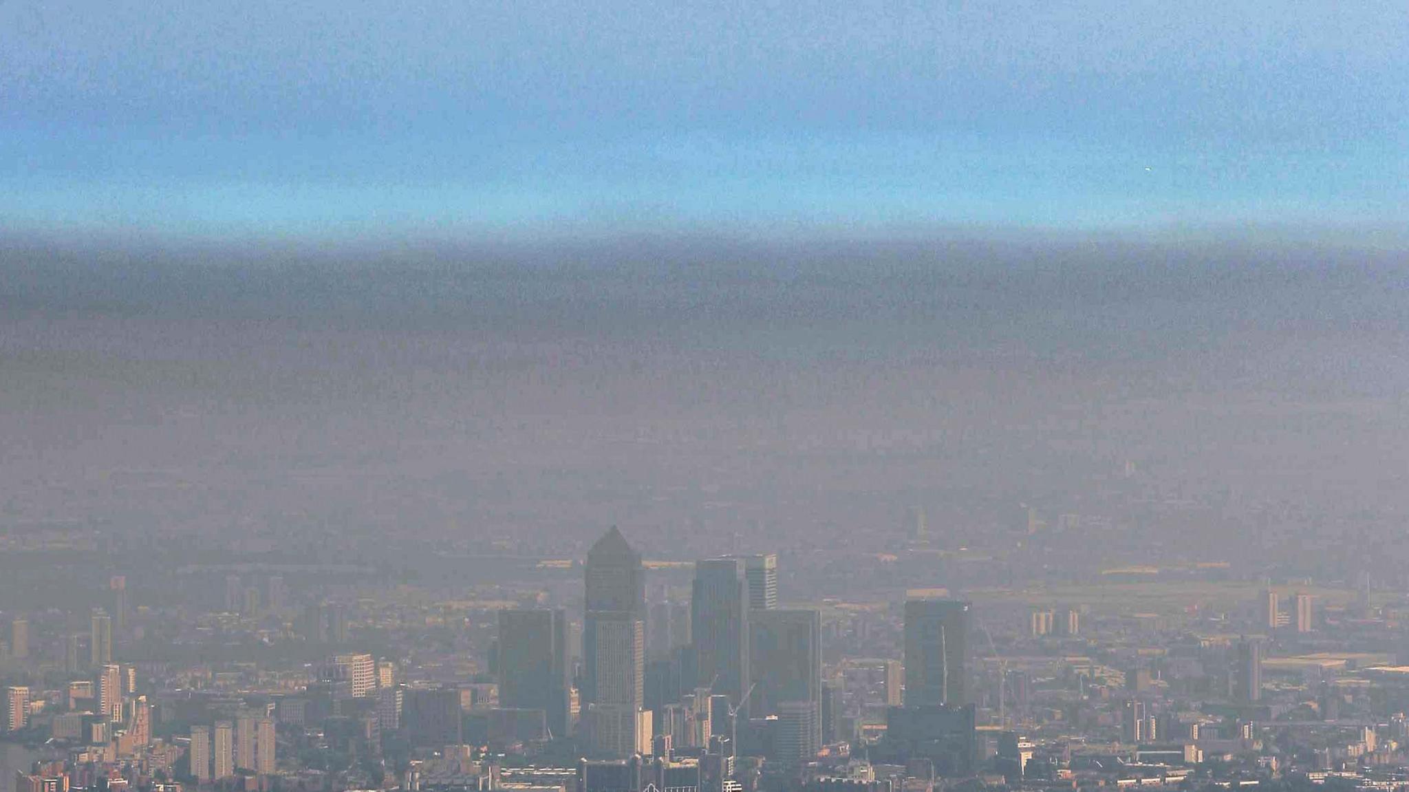 Tower blocks and buildings of London are shrouded in a grey-brown haze. The haze has a defined line two thirds up the photo with blue skies above