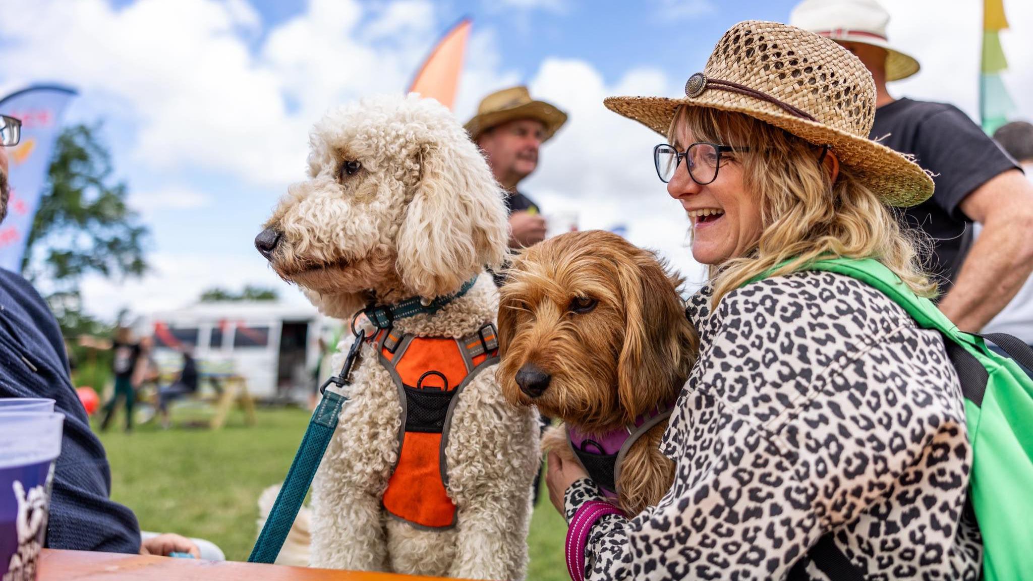 Two dogs sitting at a bench near a woman in a leopard print top who is holding one of them and smiling