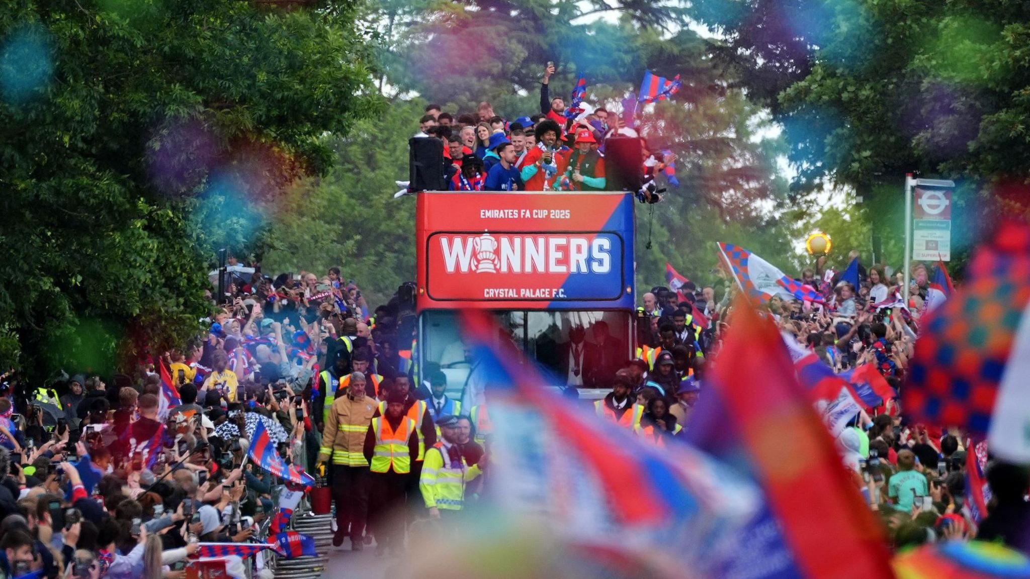 Crystal Palace players can be seen aboard the top deck of an open top bus, surrounded by crowds of fans, many waving red and blue flags