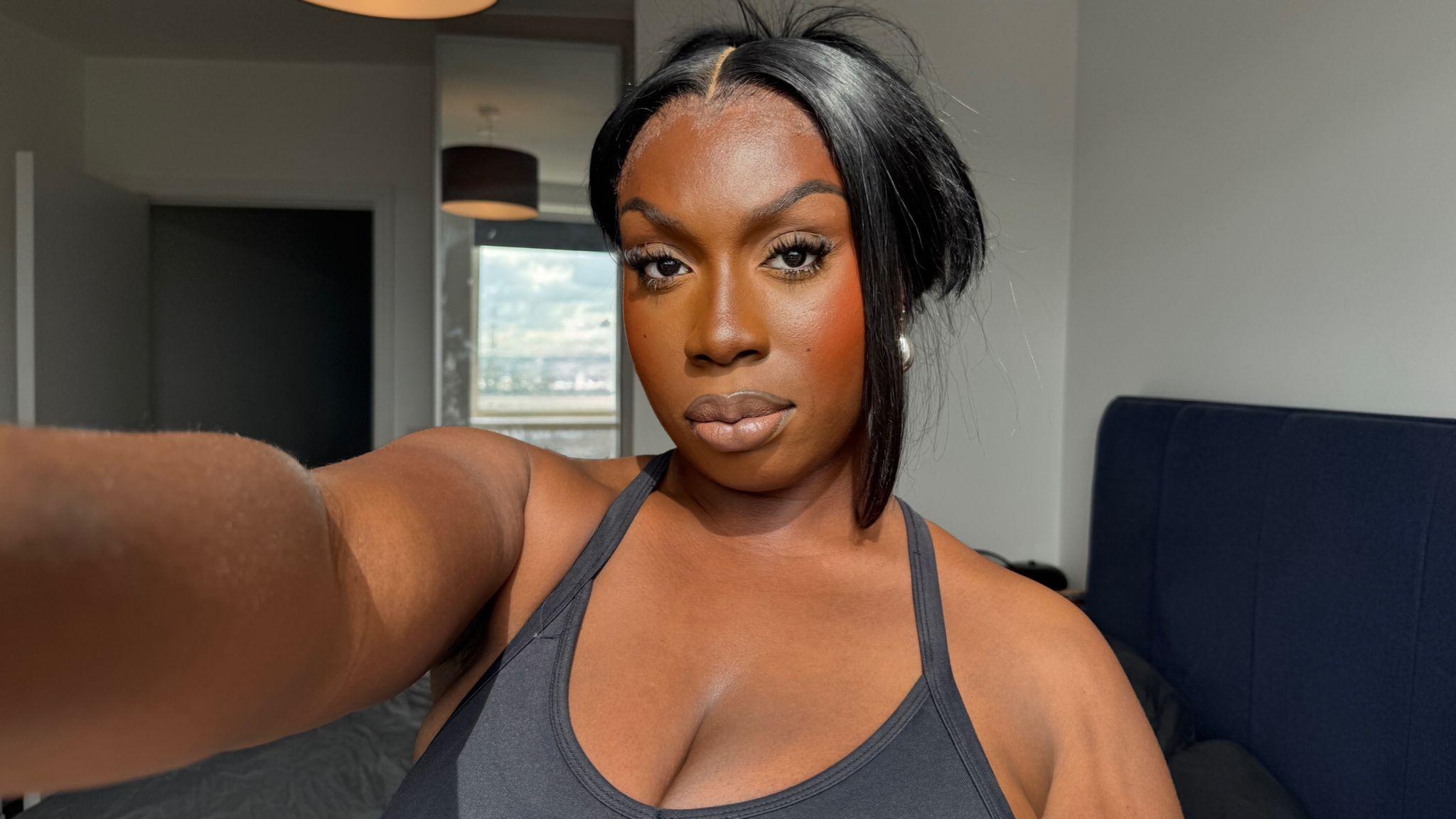 A woman in a vest top with her hair tied back holds the camera out in front of her with her right hand for a selfie. She's inside a living room with white walls and blue furniture.