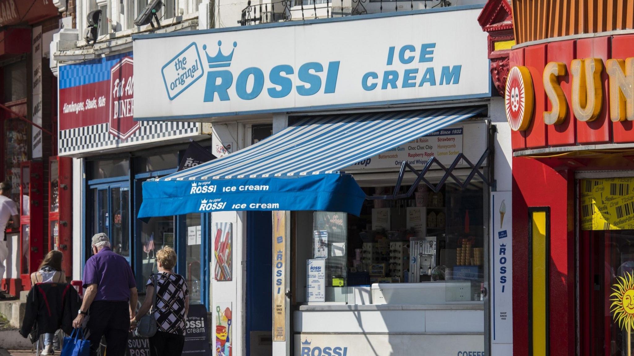 Three people walk past a shop selling ice cream. A blue and white sign says "Rossi ice cream"