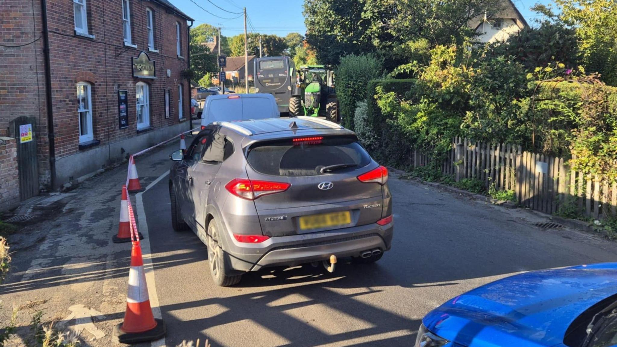 A car and a van follow a bus through a narrow road where a tractor is approaching the oncoming traffic. Cones form a make-shift pavement on the left hand side and houses and fences can be seen on the right.