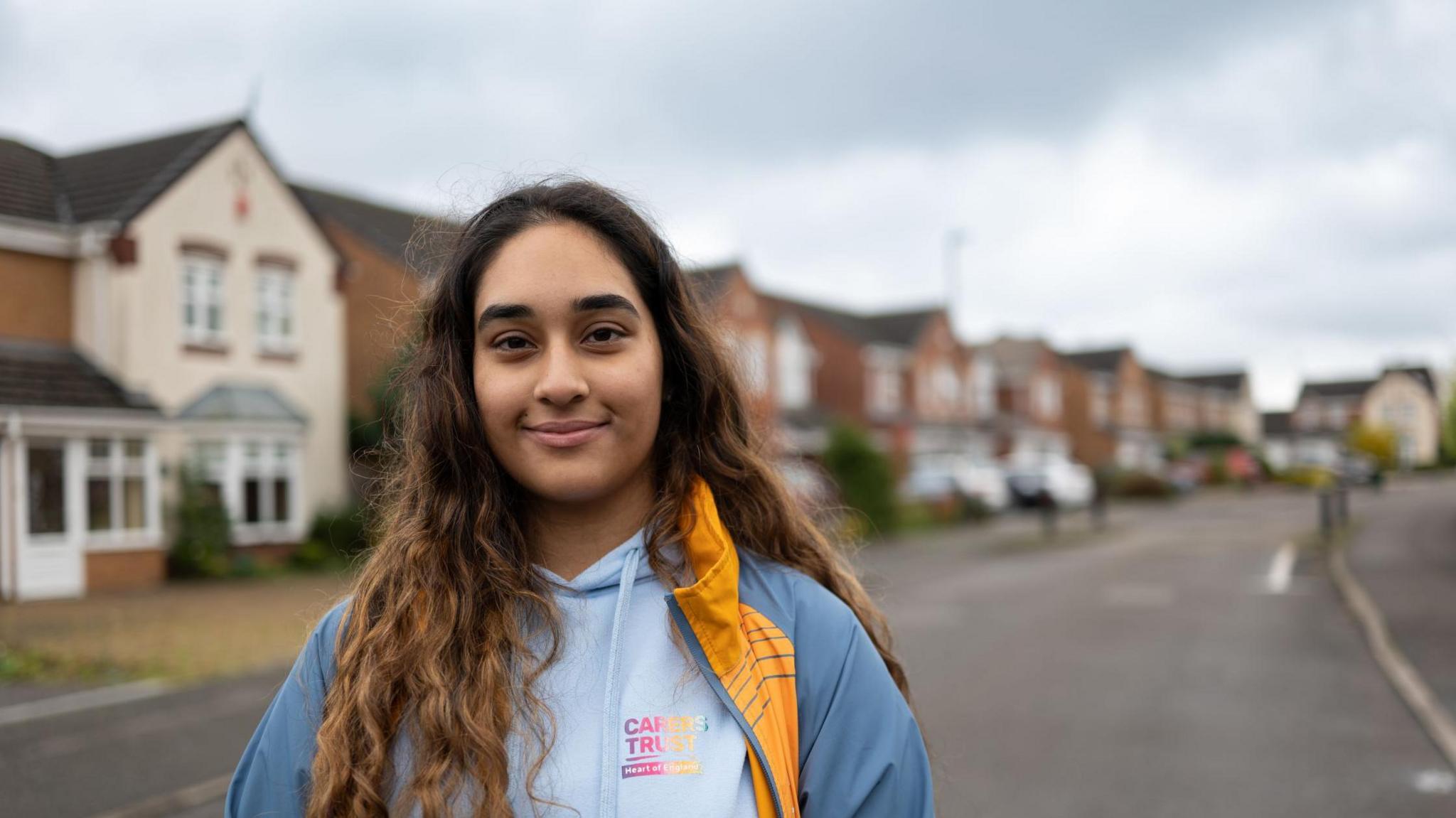 Sharandeep Sahota is a British Asian. She is standing in a residential street, wearing a blue jacket with a yellow panel, over a light blue sweatshirt from the Carers Trust.