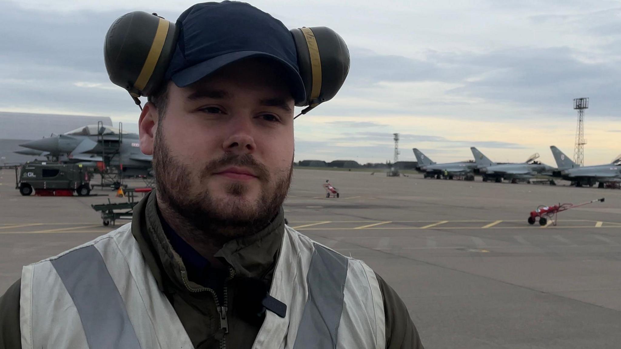 A bearded man stands on a tarmac area of an airfield. He is wearing a dark blue cap and ear defenders as well as a green jacket and high-visibility vest. Four RAF Typhoon aircraft are visible in the background.