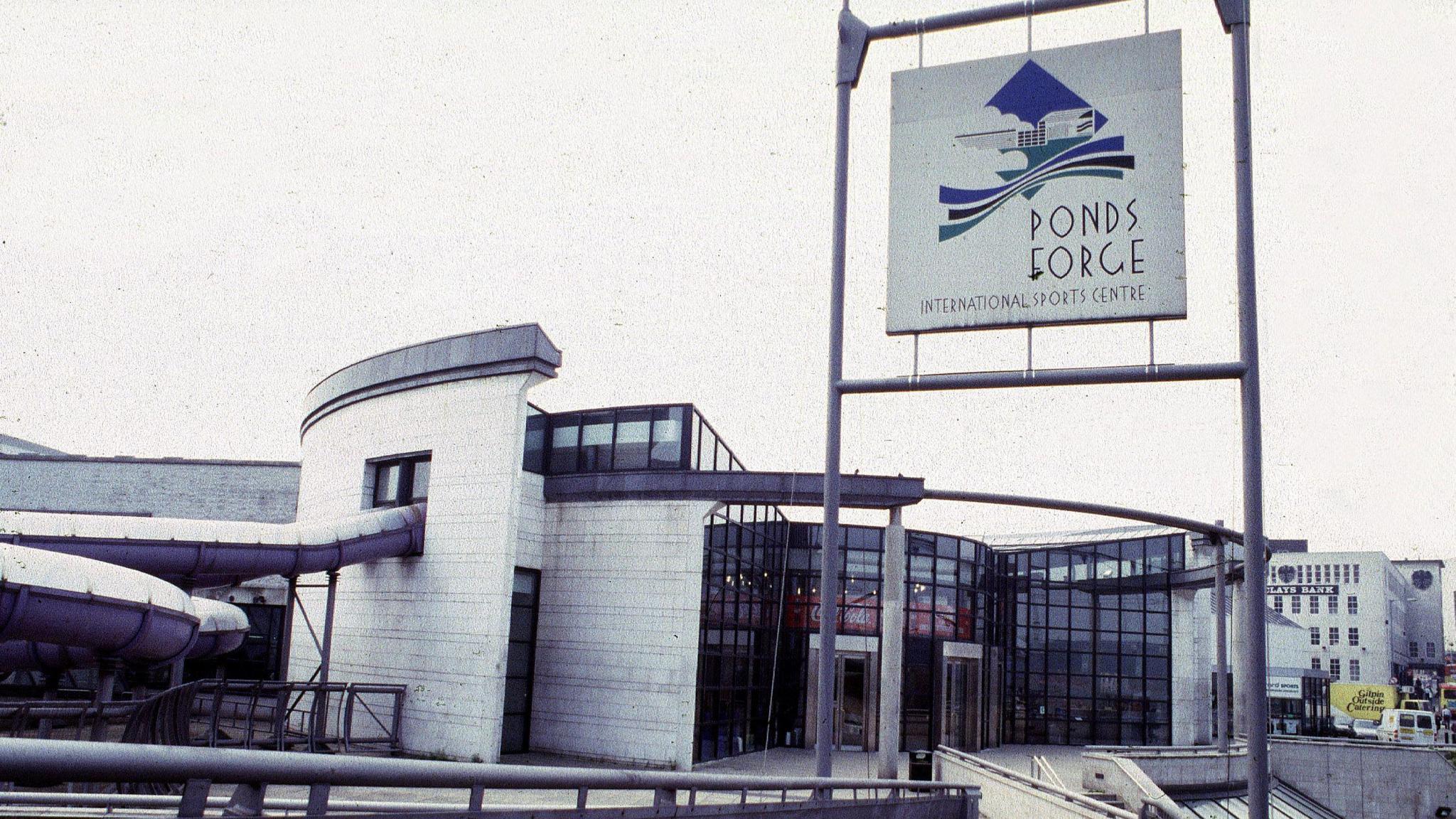 The entrance to Ponds Forge International Sports Centre in Sheffield is glass-fronted with white brick on either side. A purple water slide snakes out of the building and back in again, on the left-hand side of the image.