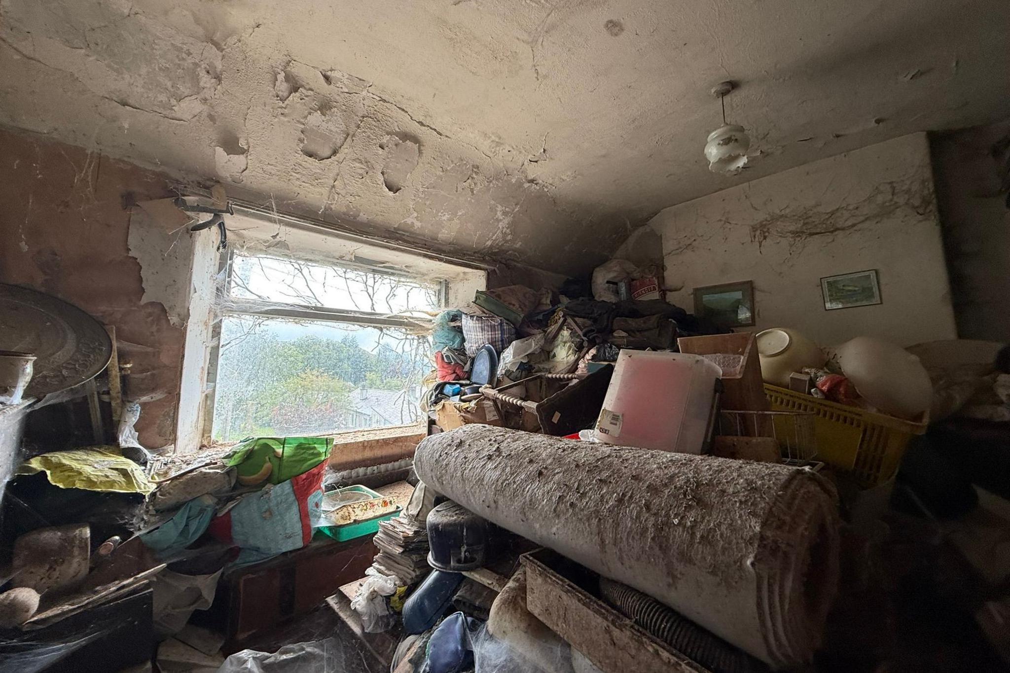 The image shows a bedroom covered in junk and dirt. Cobwebs cover the window and walls. The junk covers the majority of the room.  A large rug is at the centre of the room.