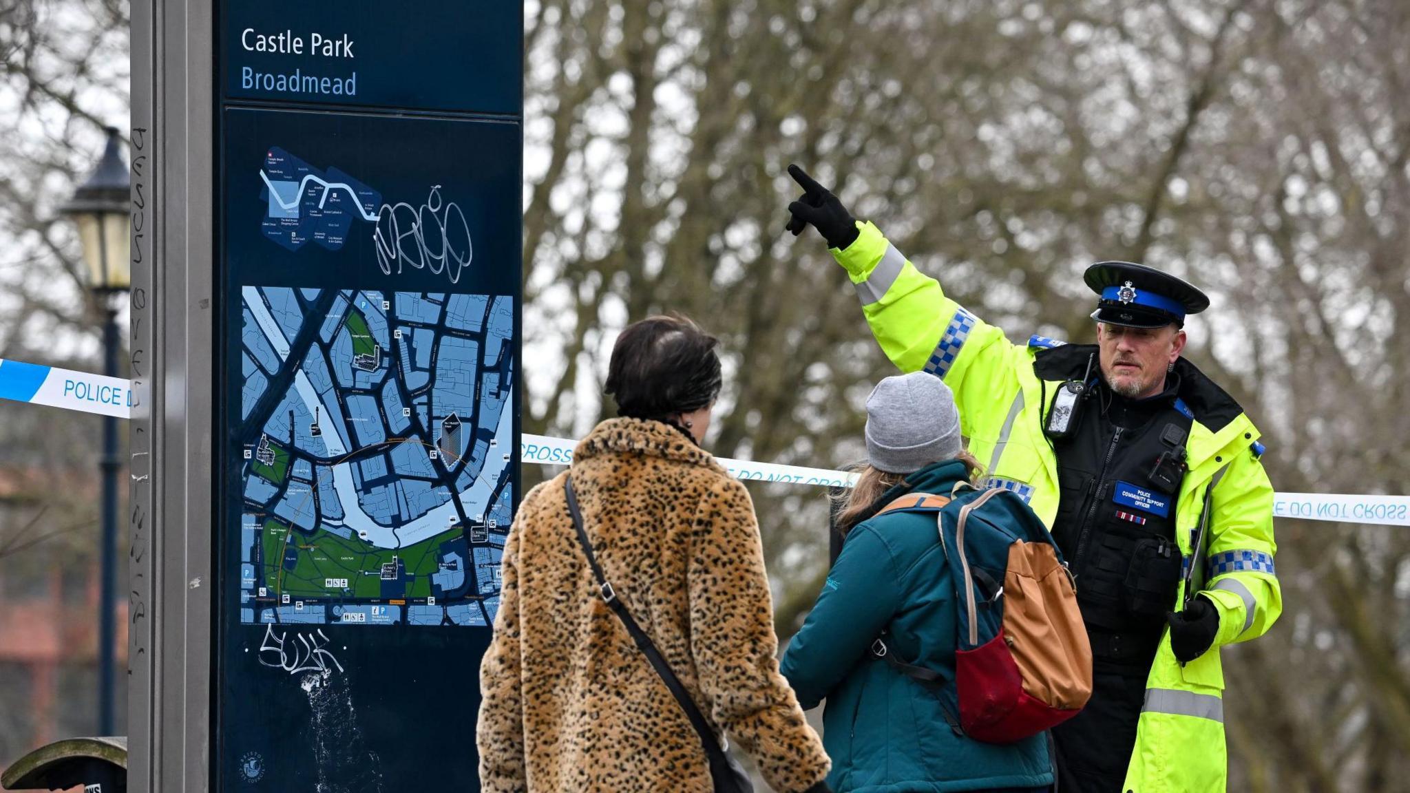 A police officer wearing a high-viz jacket over his uniform directs two women with his arm as he stands in front of a blue and white police cordon next to Castle Park in Bristol city centre. A sign with a map of park behind him