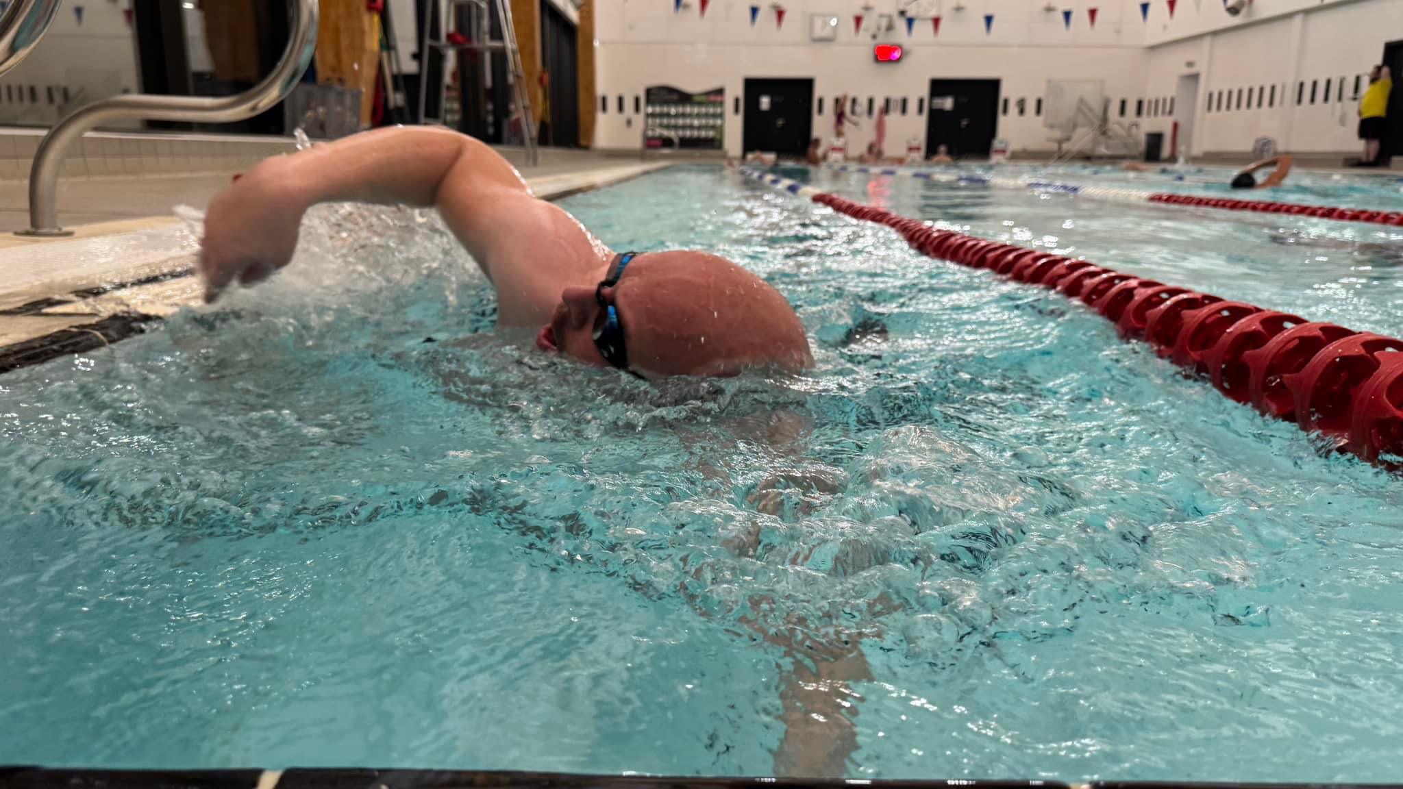 A man is swimming in a swimming pool. He is wearing black goggles, and there is a red lane divider next to him. He is swimming the front crawl and has one arm out of the water