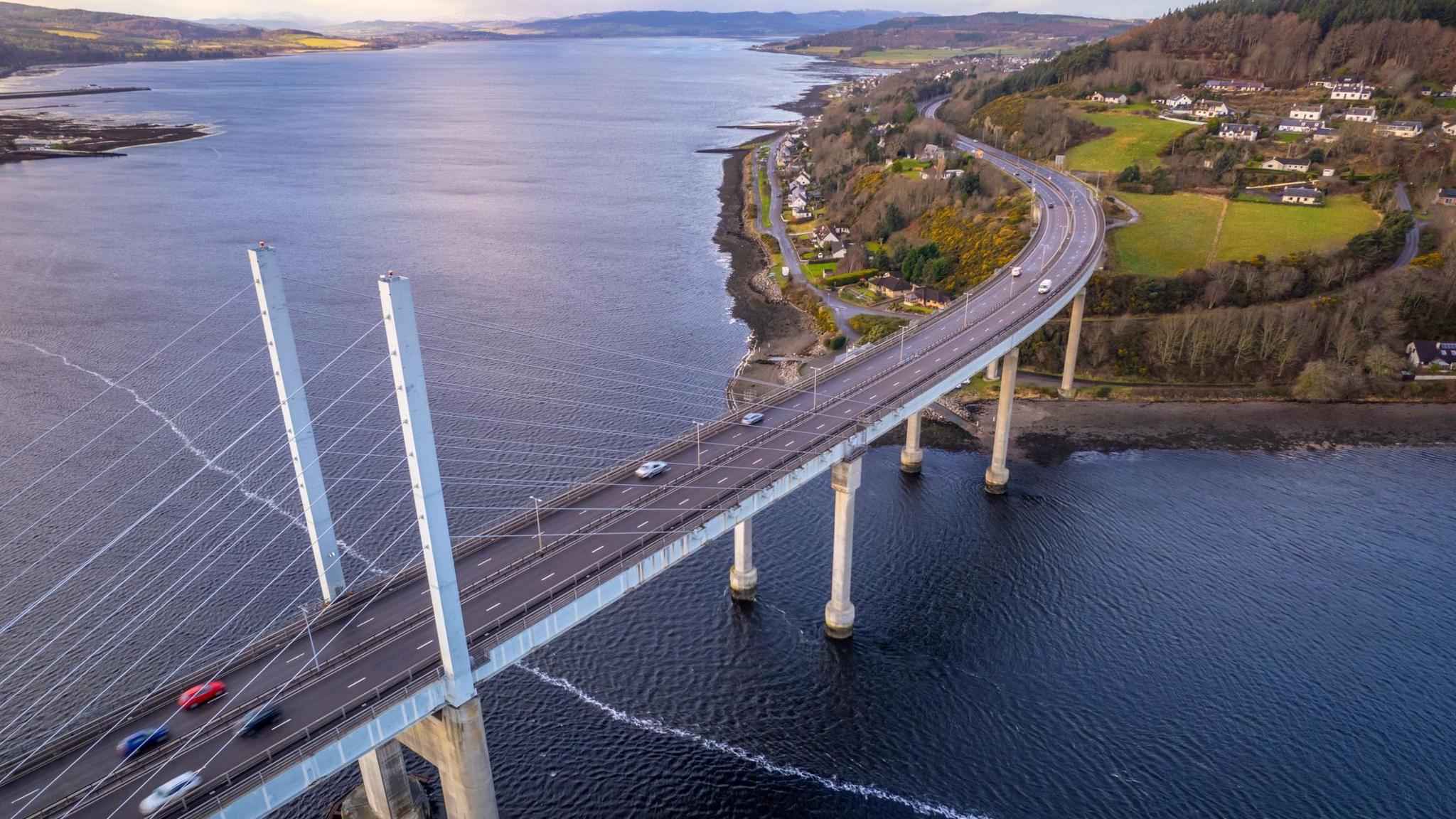 An aerial view of the bridge. There are cars on the carriageway. The water that is visible is of the Beauly Firth, on the left, and Moray Firth to the right of the bridge. Underneath the crossing is the Kessock Channel.