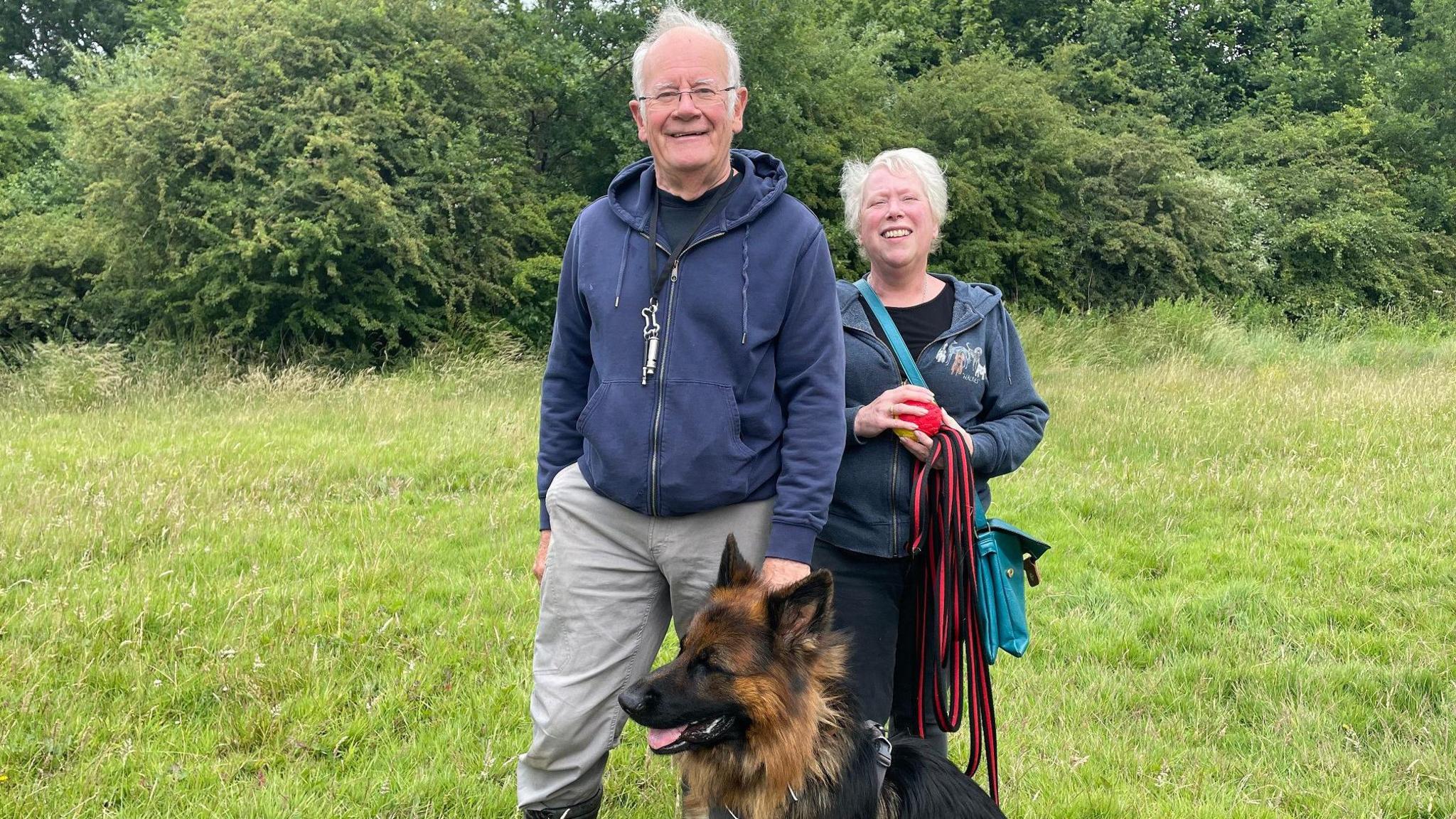 Two dog walkers and their pup stood in a green space