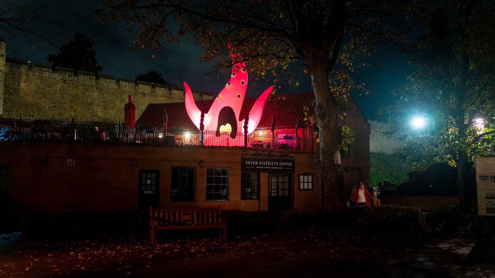 A lit-up inflatable pink monster can be seen flailing its arms in an outdoor seating area at the top of a restaurant. It has a long pointy head and arms with several eye balls and a mouth pulling a scared face. It is night time and the front of the restaurant reads 'Seven Districts Coffee'.