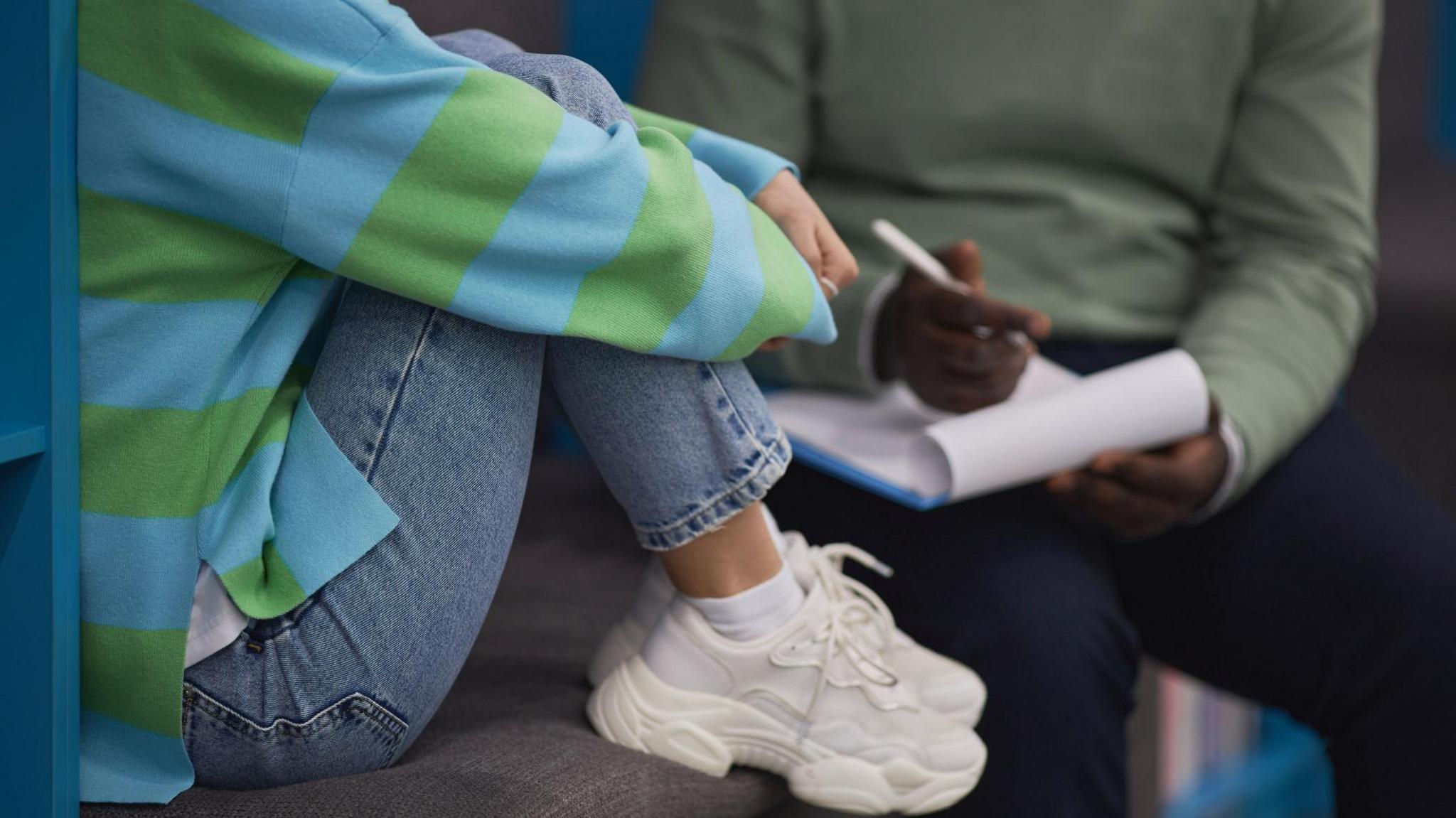 An unidentifiable child curled up on a seat speaks to an adult holding a notebook