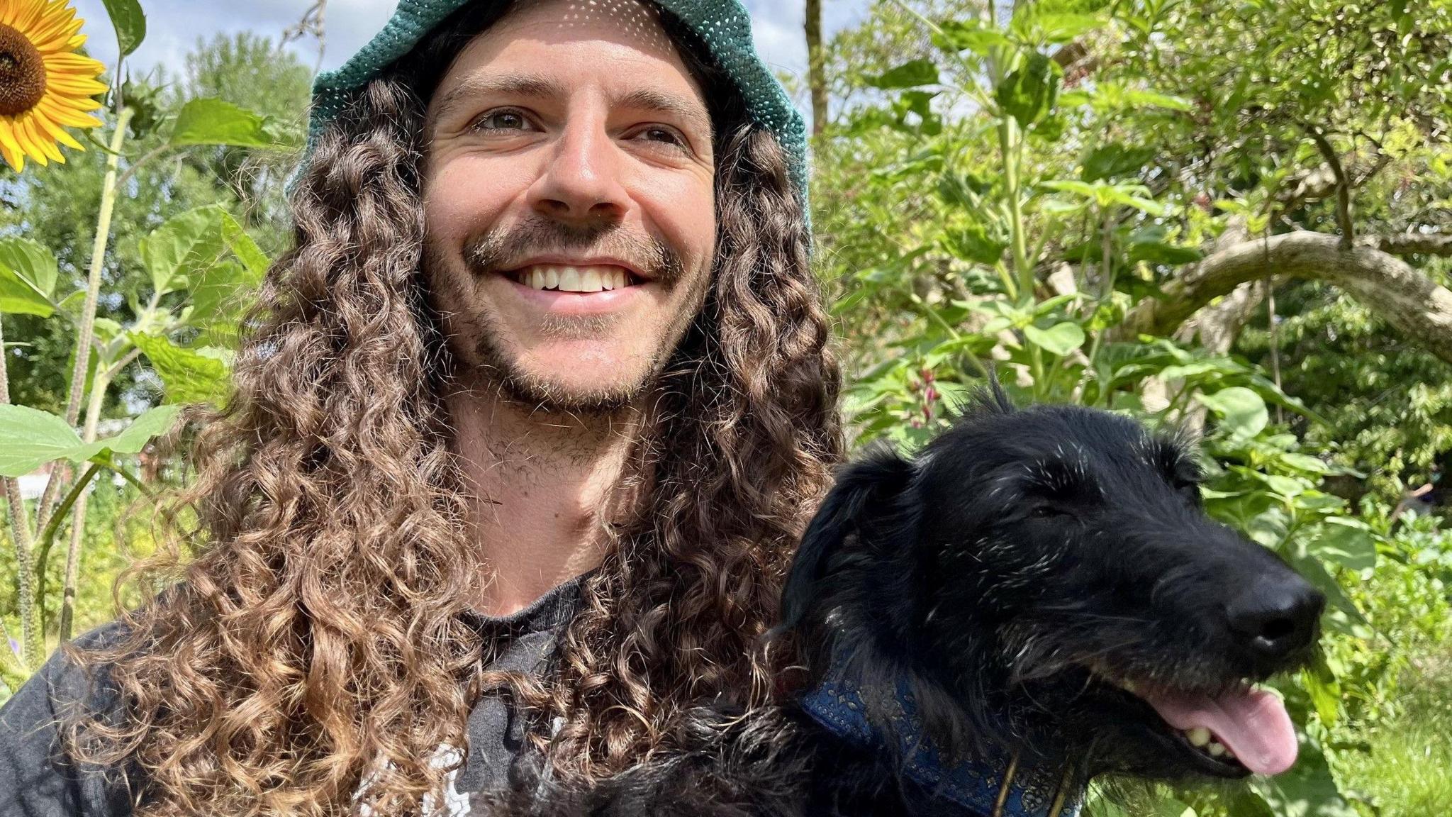 A man with dark, long curly hair holds a small black dog, with green plants in the background