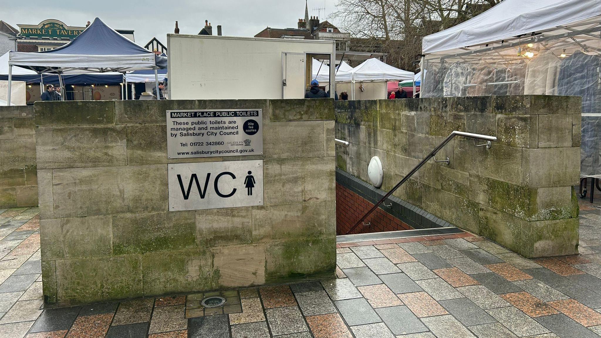 Market Square toilets in Salisbury. A staircase leads down to the block which is underground. There is a wall around the top of the block and a WC sign on the side. Market stalls can be seen in the distance.