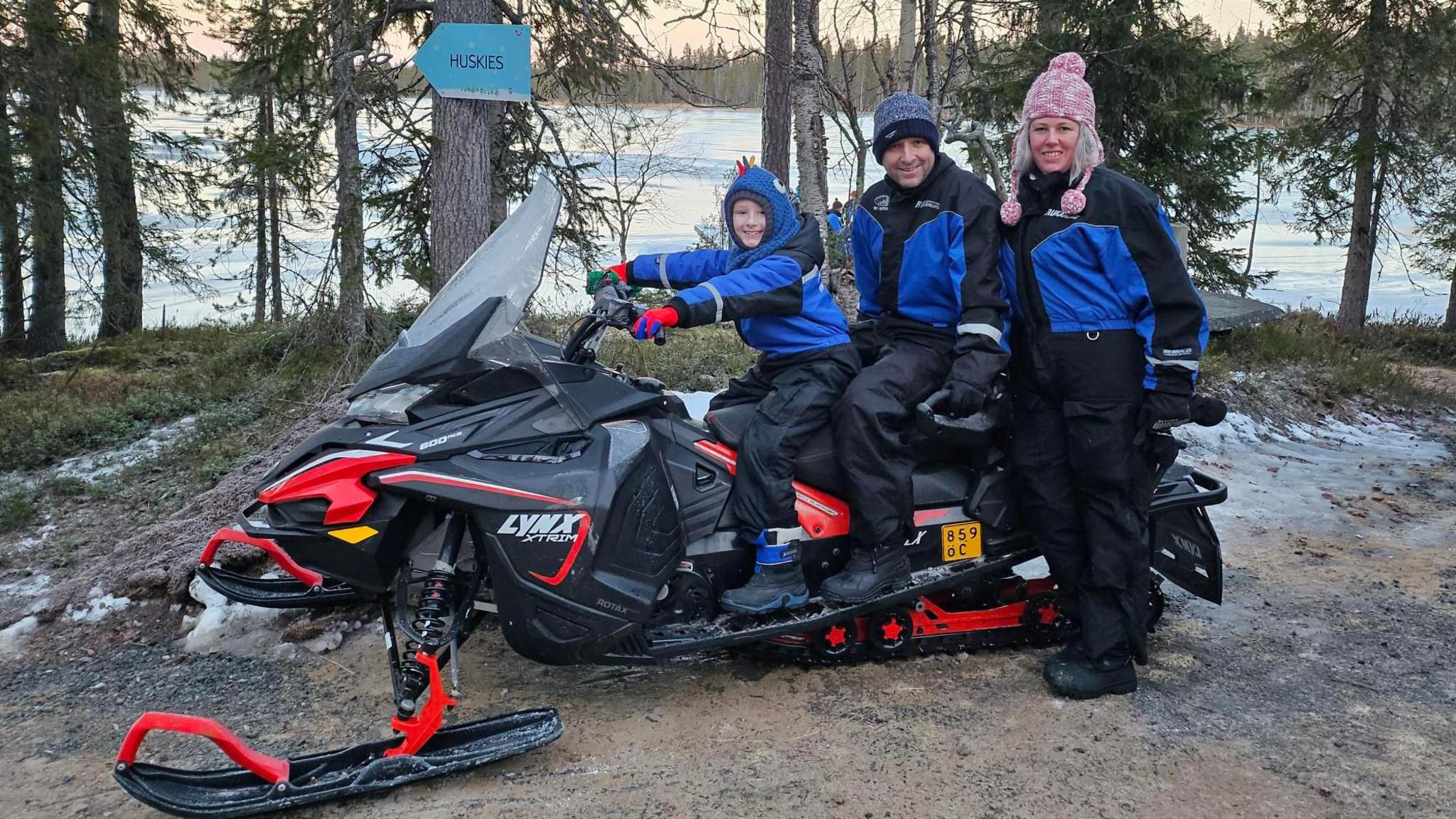 Wayne O'Neil and his son Finn sitting on a snowmobile with Lisa O'Neil standing next to them. They are all wearing snow suits and woolly hats, but there is no snow on the ground around them. There is a lake in the background.