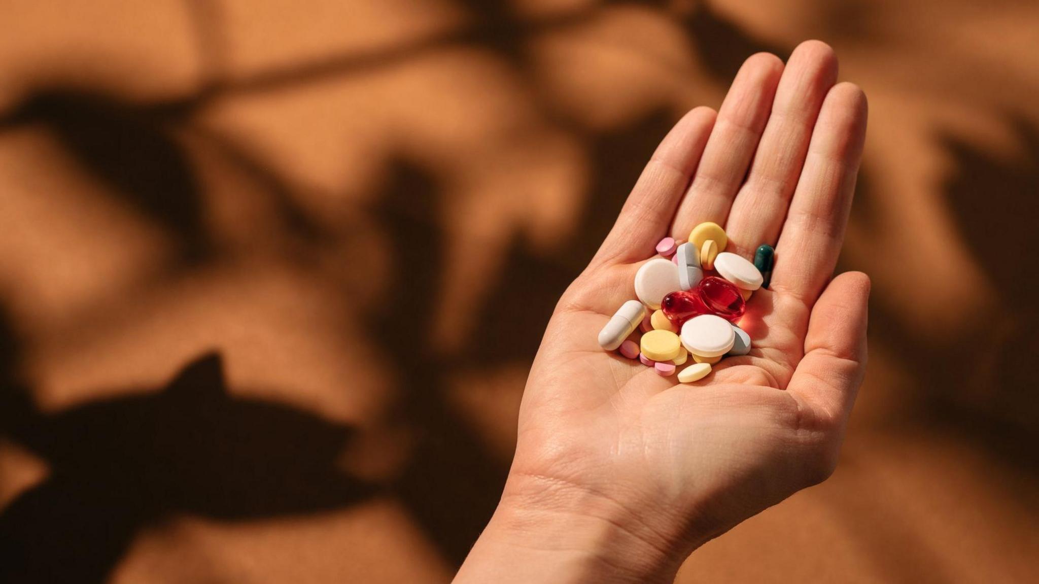 Woman's hand holding colorful pills against terracotta table background with shadows from home plant. Pile of different pills - white, red, yellow, blue, green at the hand. 