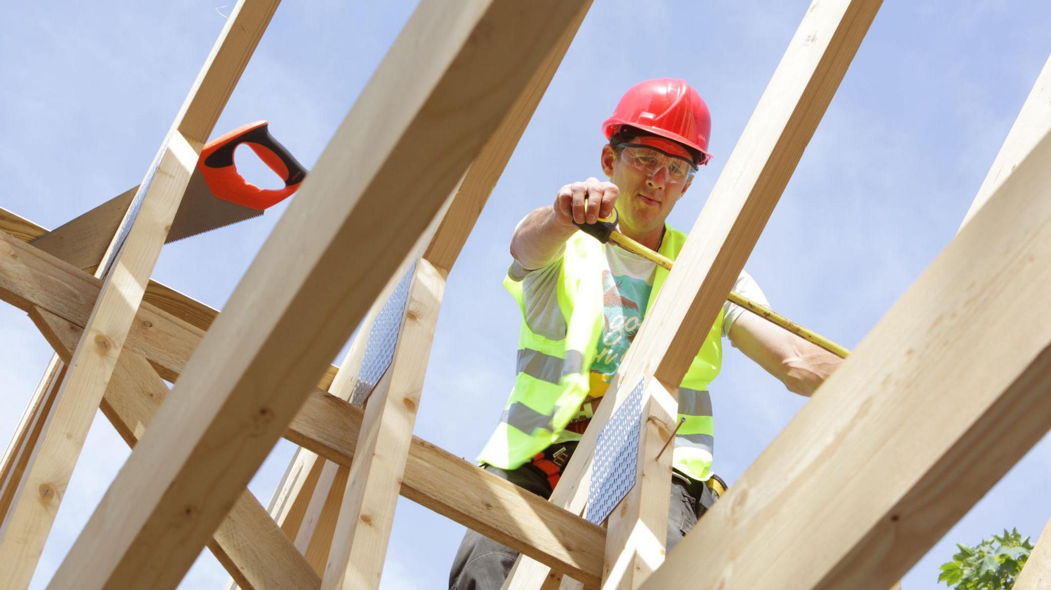 A man working on the roof of the house - the shot is taken looking up towards him from inside the house as he works on building its roof. A series of pale wooden beams criss-cross the image as the man stand on them to use a measuring tape. He is wearing a red hard hat, high-viz jacket and grey trousers.