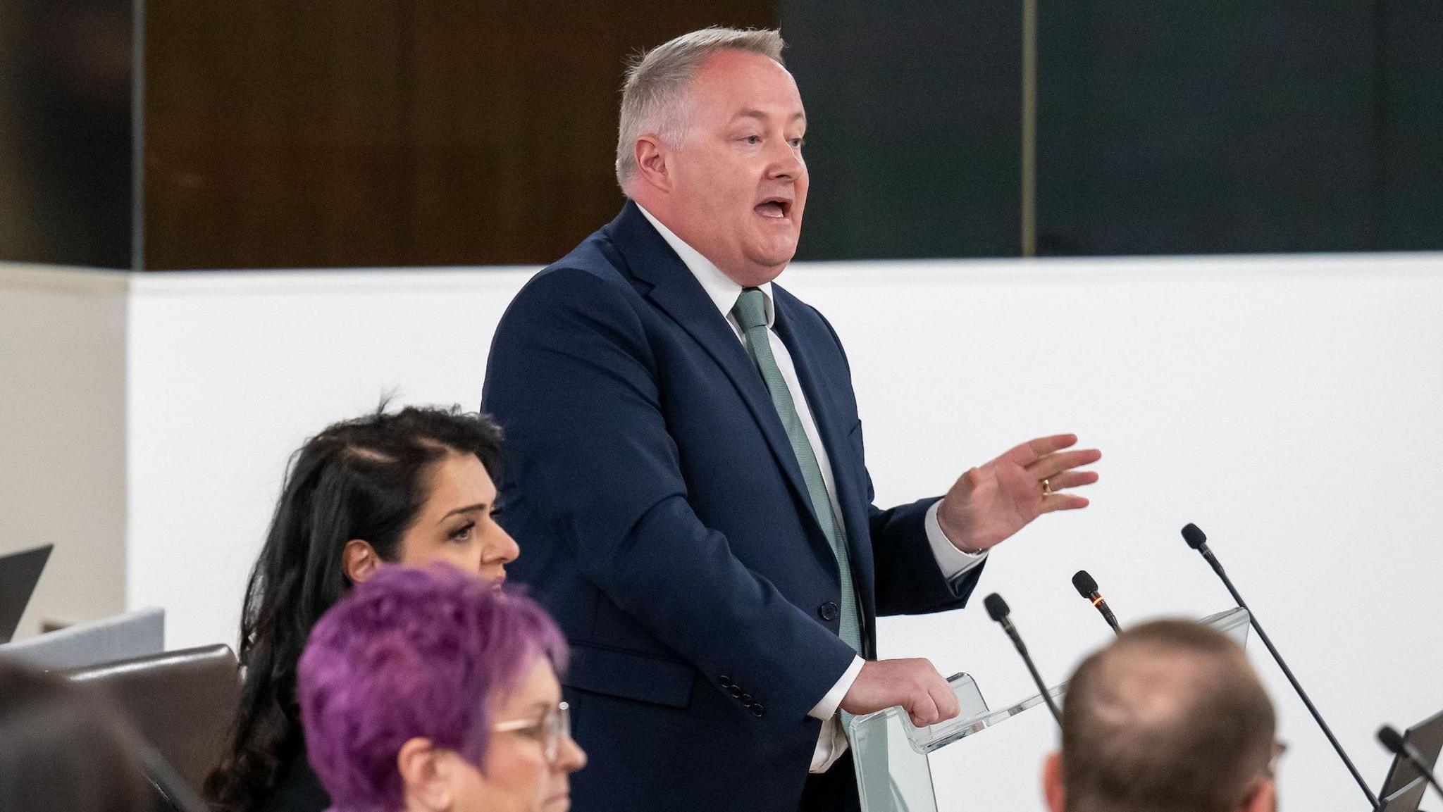 Darren Millar stood speaking in the Ty Hywel debating chamber, wearing a blue suit, with other Members of the Senedd in the foreground sat down.