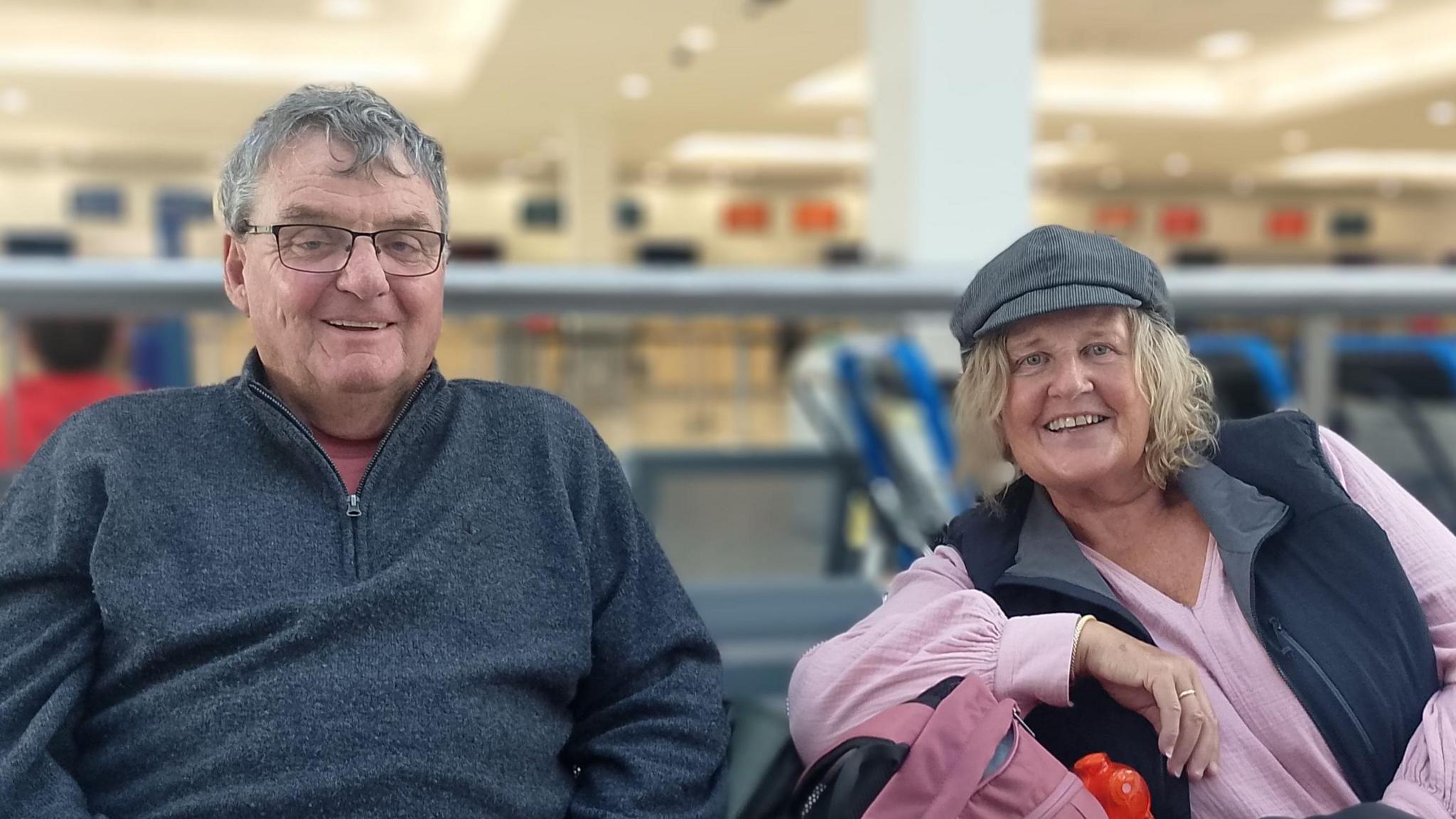 A photo inside the airport where a man and woman sit with their luggage. The man, who is wearing glasses, rests his phone on his knee. The woman is leaning on a bag pack.