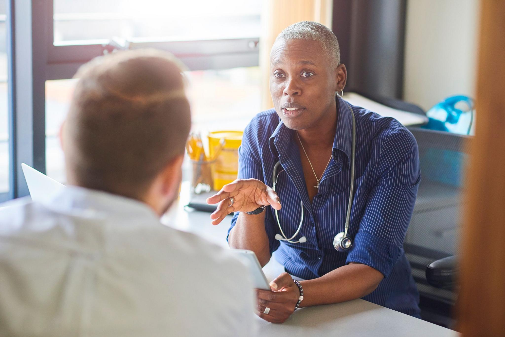 Doctor wearing blue pin-stripe shirt and stethoscope around neck, explaining something to patient who has back turned to camera. Patient is wears a white shirt and has light brown hair 