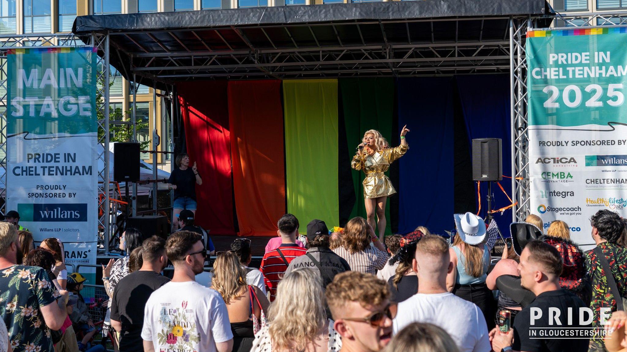 A performer dressed in a gold-coloured outfit is singing on stage while dozens of people watch. On the side of the stage is a sign which reads: "Pride in Cheltenham 2025".