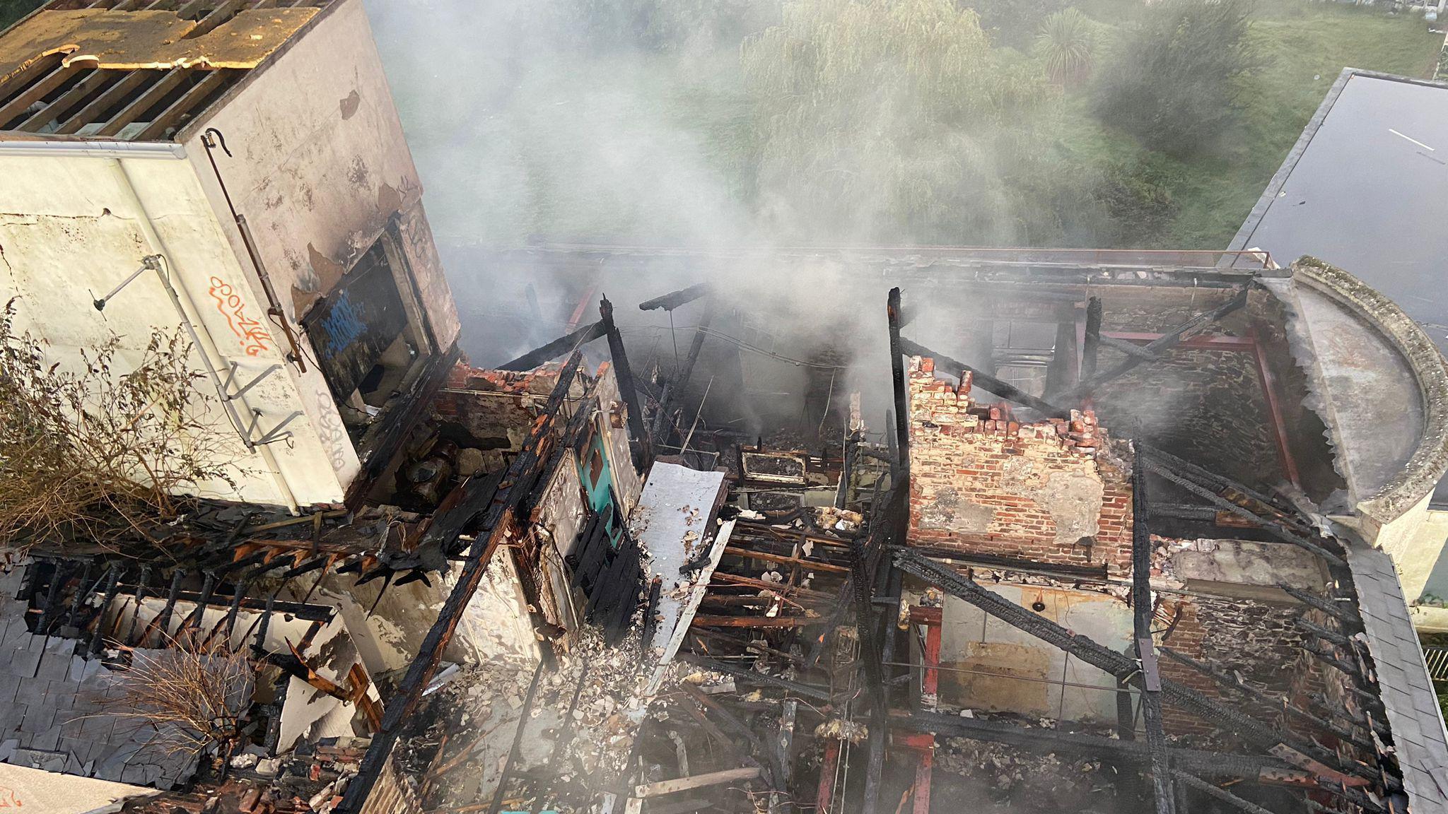 Derelict hospital building is badly damaged with smoke still visible. Walls have caved in and the remaining beams are badly burnt. 