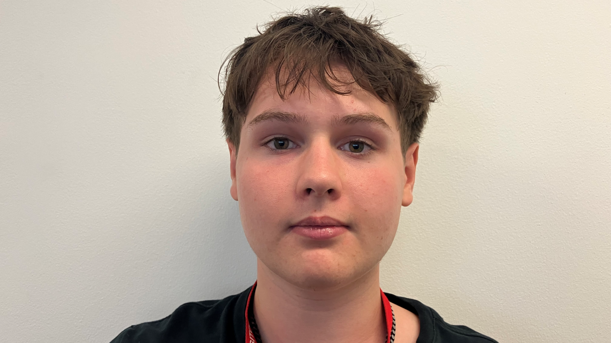 A teenage boy with dark brown hair wearing a black t-shirt and a red lanyard saying 'visitor'