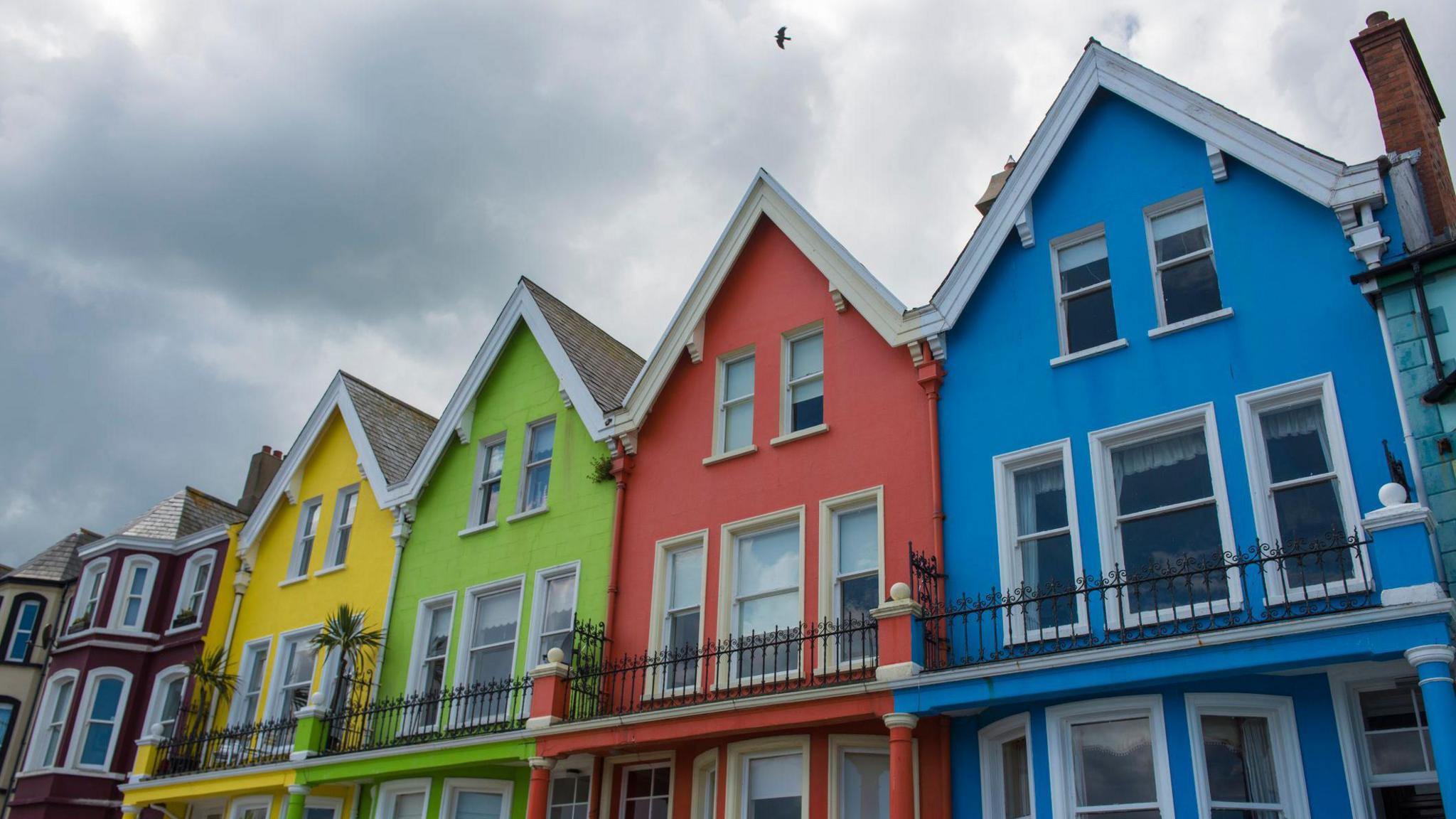 Whitehead, County Antrim. A row of terraced coloured (blue, red, green yellow and aubergine) houses on the waterfront. They all have a black metal balcony and white white frames.