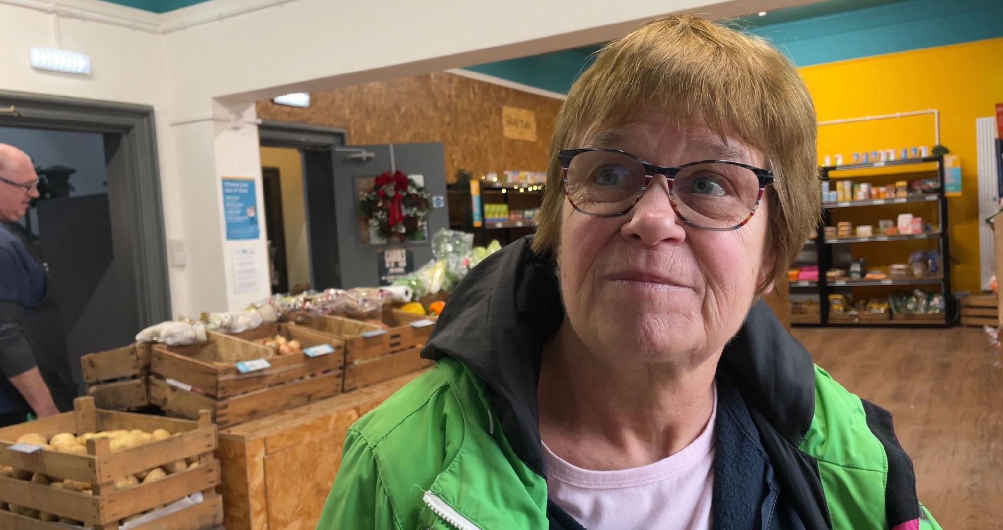 A lady wearing glasses and a green and black jacket looks up at the camera. She is standing in a grocery shop with shelves full of produce in the background