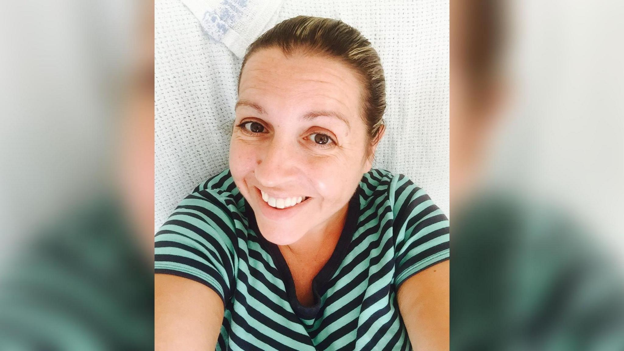 Ali Alcock is smiling in this selfie taken as she lies on a hospital bed. She is wearing a mint green and blue striped T-shirt.