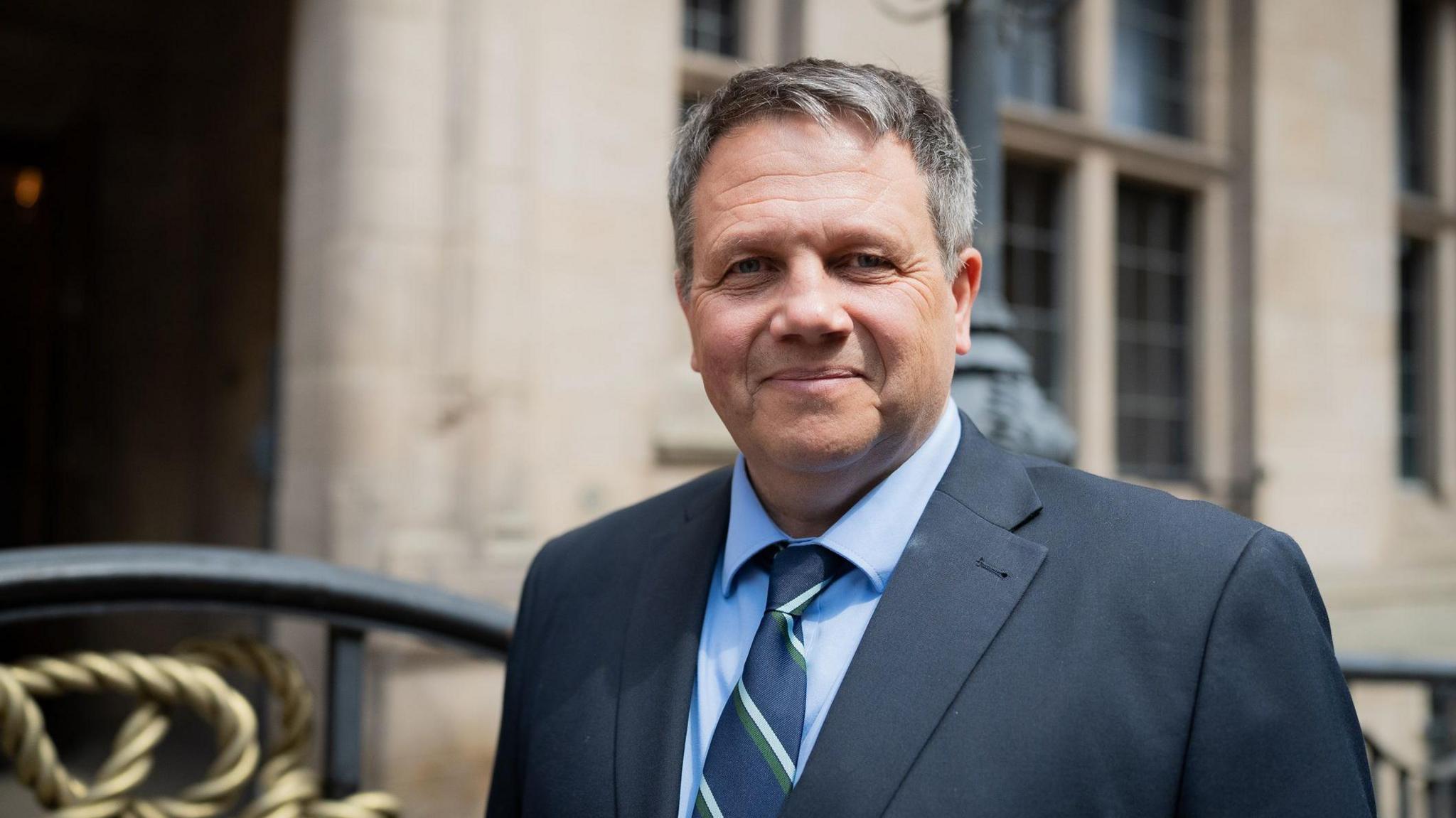 Andrew Mynors, a man wearing a dark suit with a blue shirt and striped tie. He is standing outside the County Buildings in Stafford, a