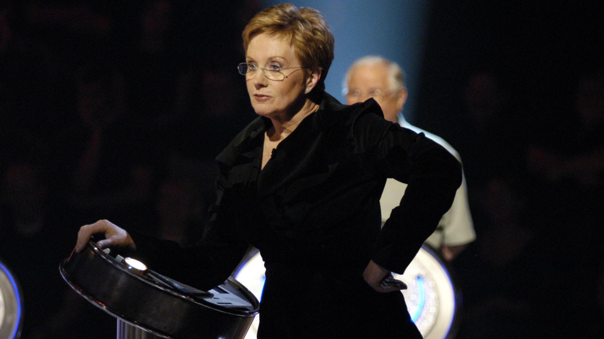 A younger Anne Robinson, dressed in black, with one hand on her hip and one hand on her presenting panel