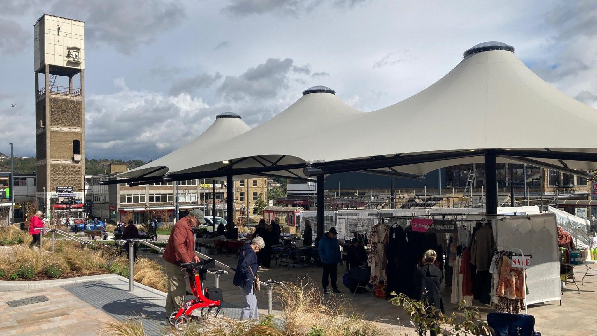 A collection of market stalls under a white canopy with a concrete clock tower in the background.