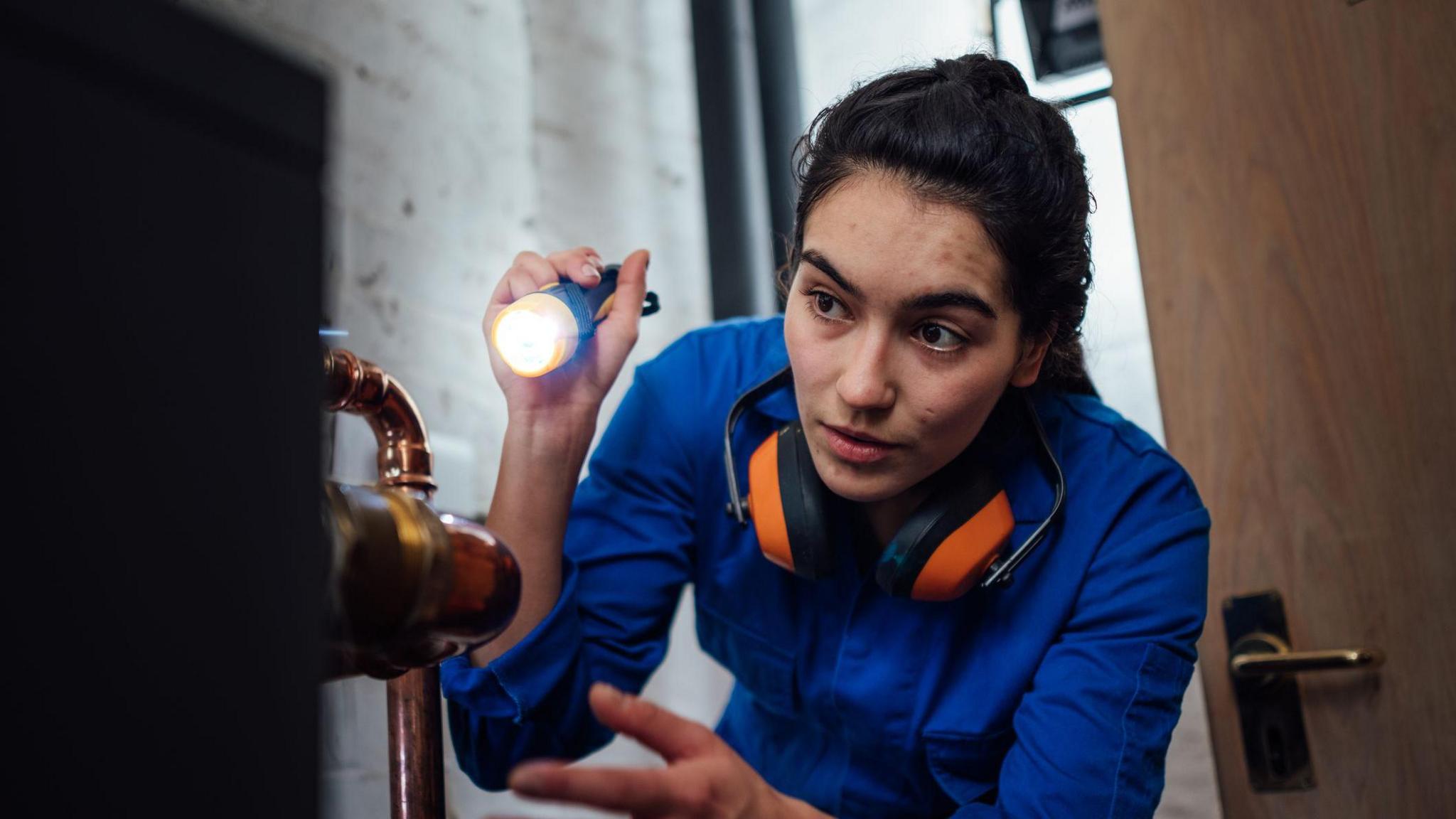 Young woman in boiler suit with ear defenders around her neck looking with a torch at a boiler