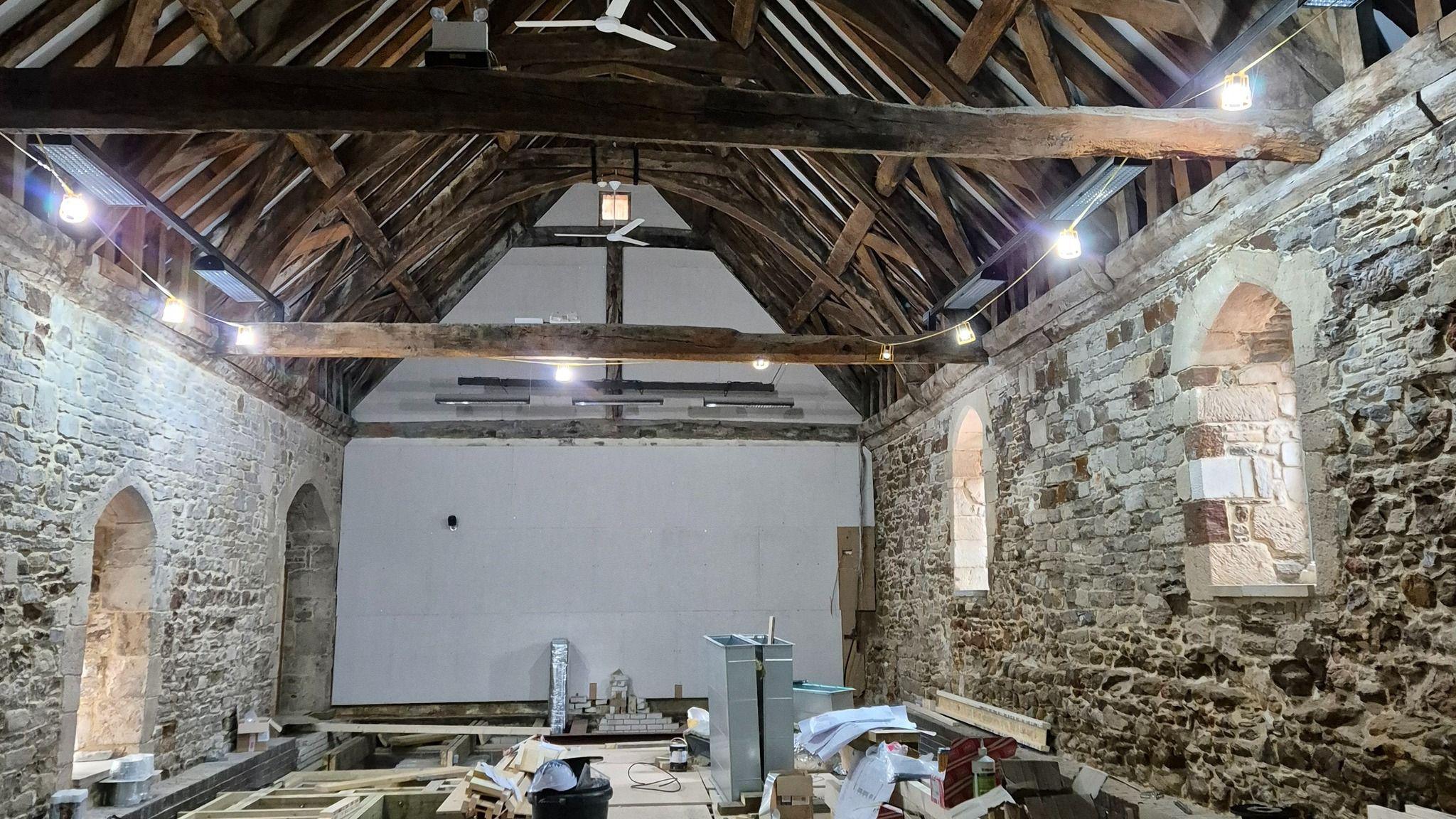 The interior of a medieval hall with stone walls and a pitched wooden roof. The floor is under construction and the area is covered with tools and components