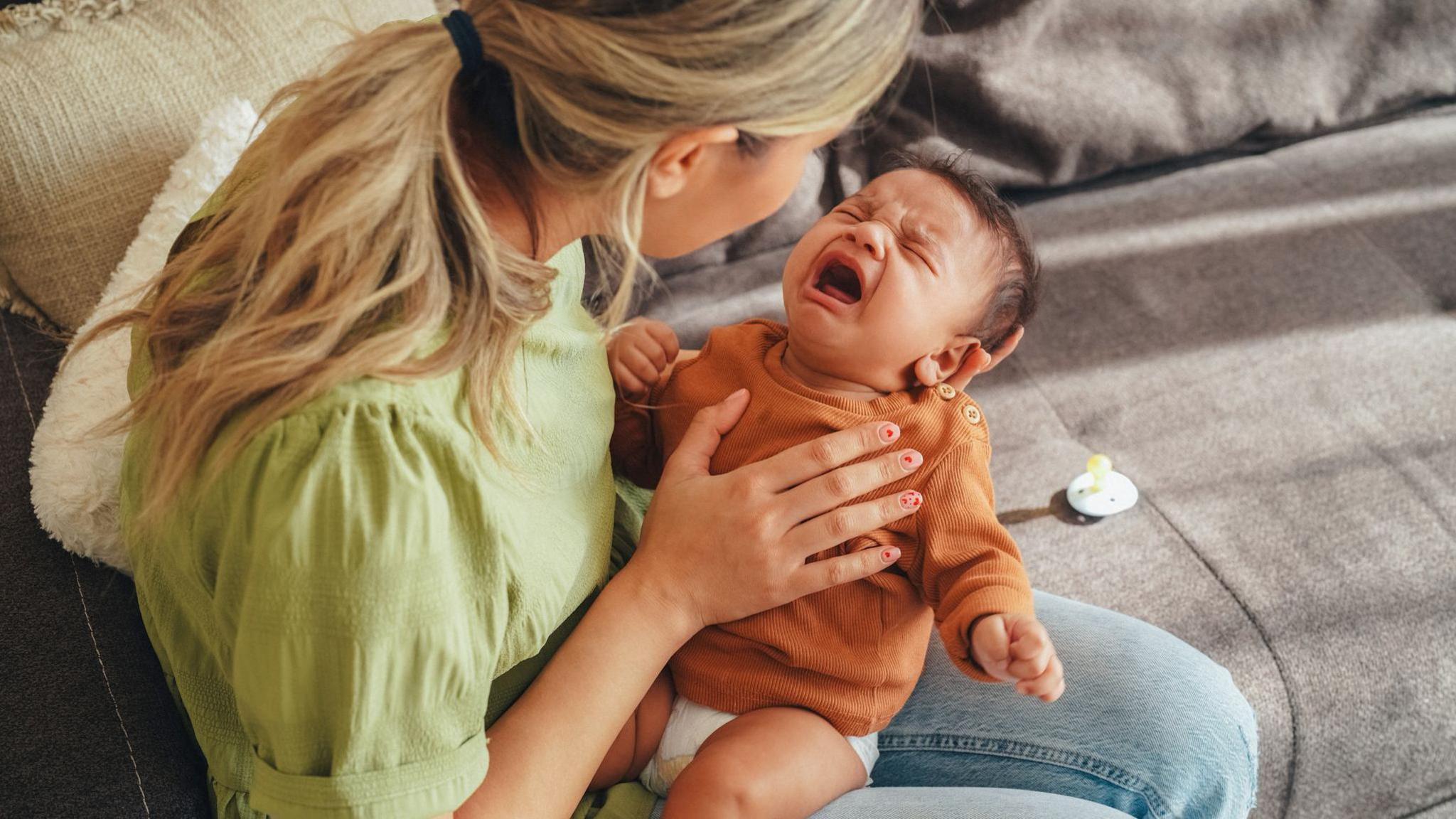 A mother with blonde hair wearing a light green blouse holding her crying baby on her lap. She has one hand on the baby's chest and the other holding  up it's head. They are sitting both on a grey sofa and the baby's dummy is laying beside them.